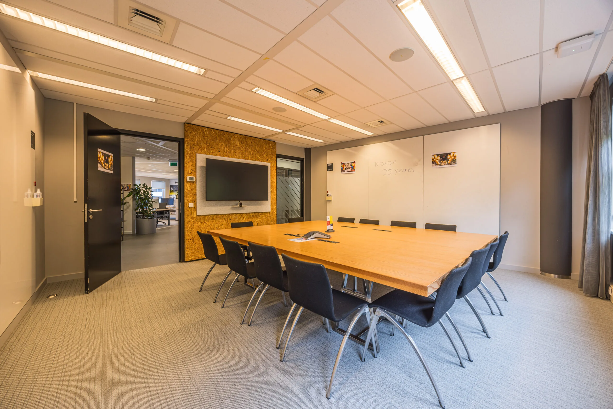 Modern conference room with a large wooden table, black chairs, wall-mounted screen, and whiteboard reading "Kidota 25 years".