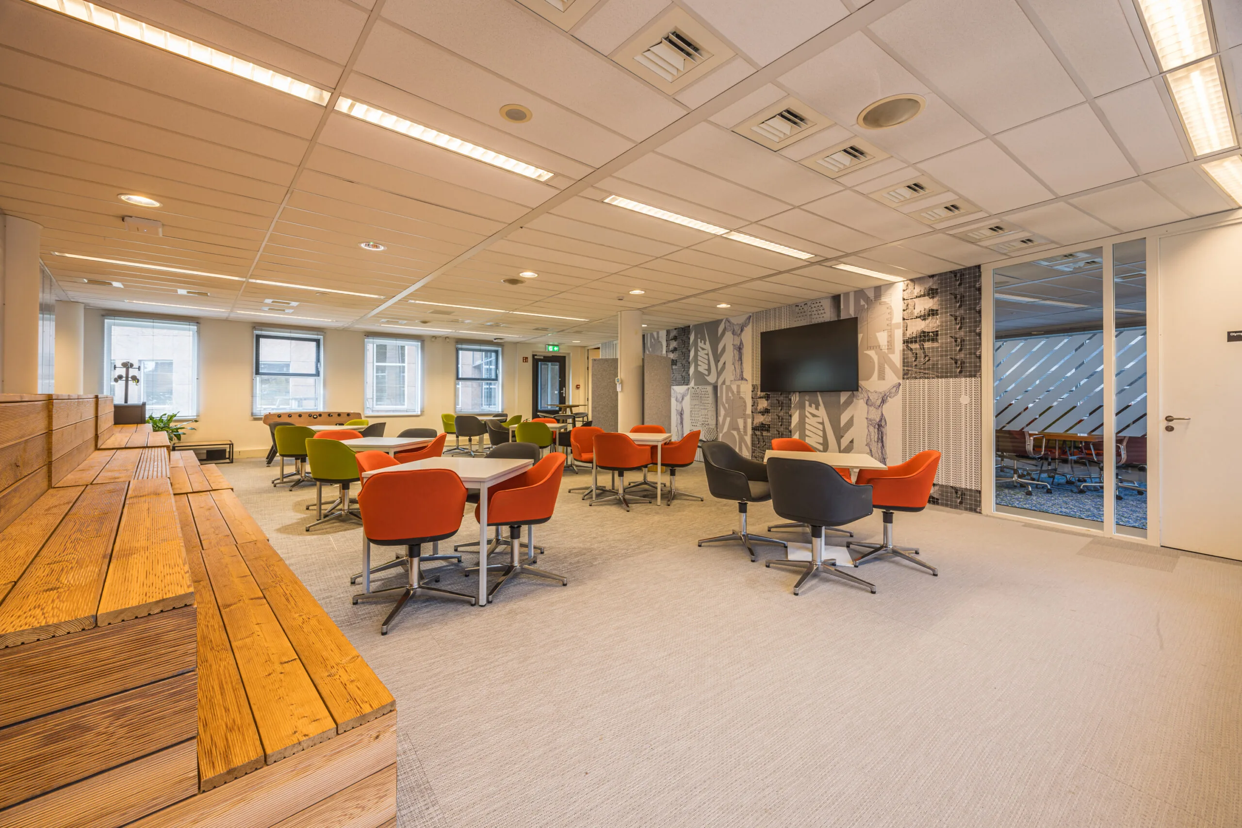 Modern office lounge with colorful chairs, wooden seating steps, and a wall-mounted screen in the Olympia workspace.