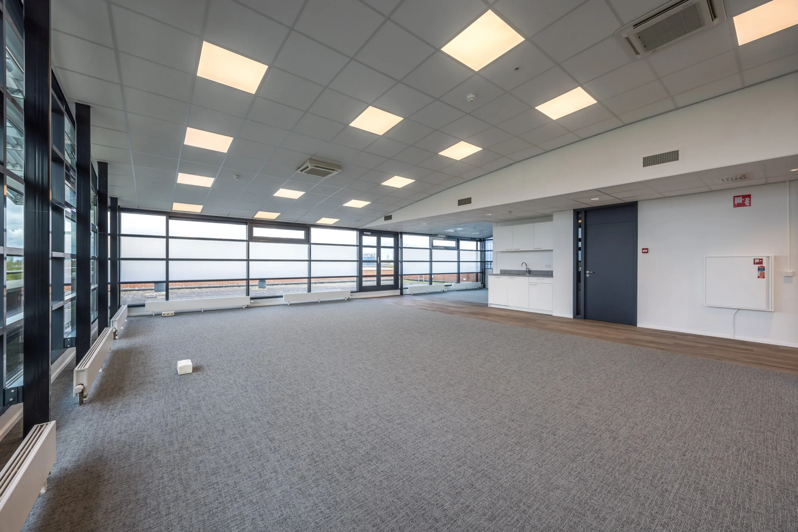Modern empty office space at Veldzigt with large windows, carpeted floor, and a small kitchenette.