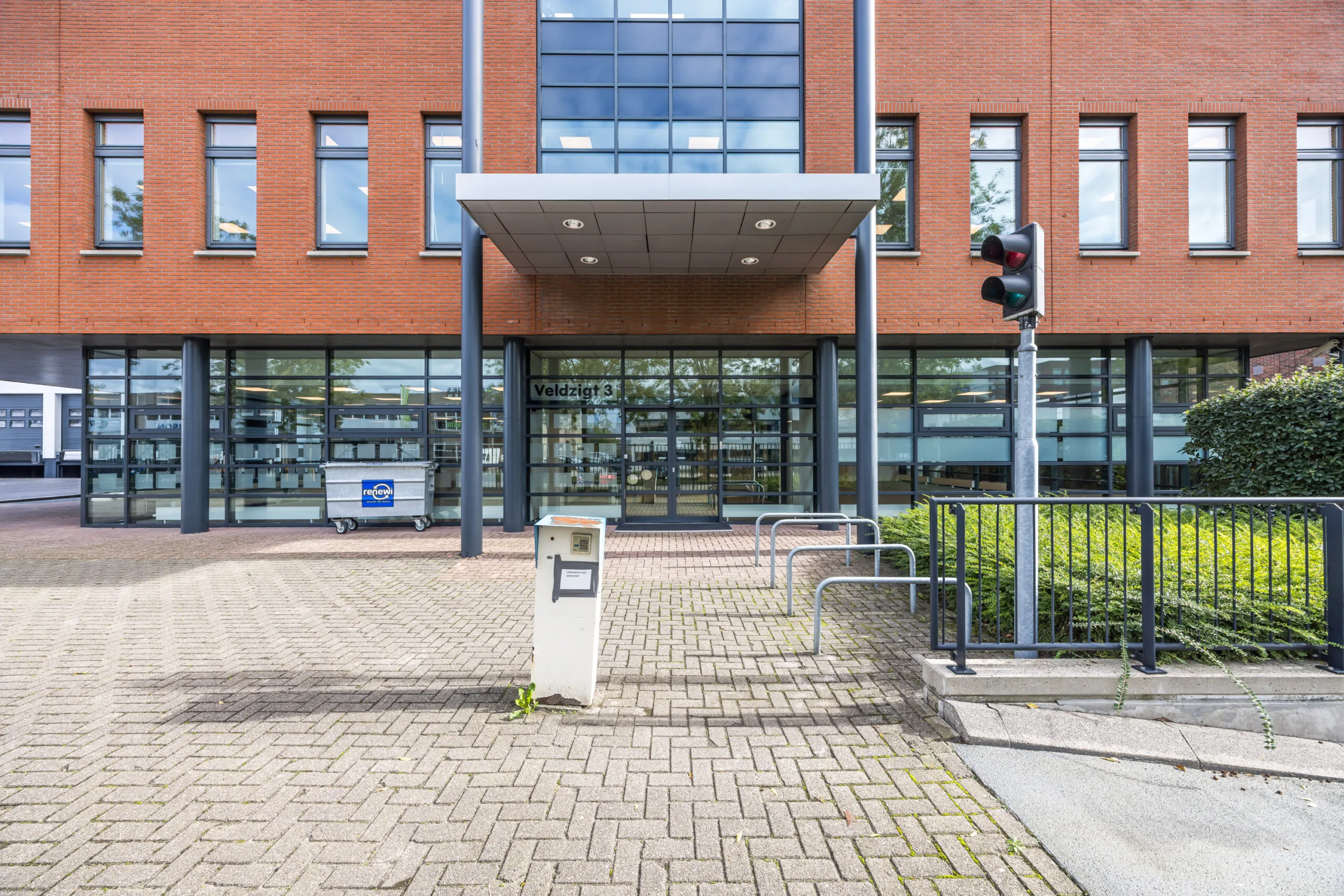Entrance of the Veldzigt 3 office building with bicycle racks, a traffic light, and a recycling container in front.