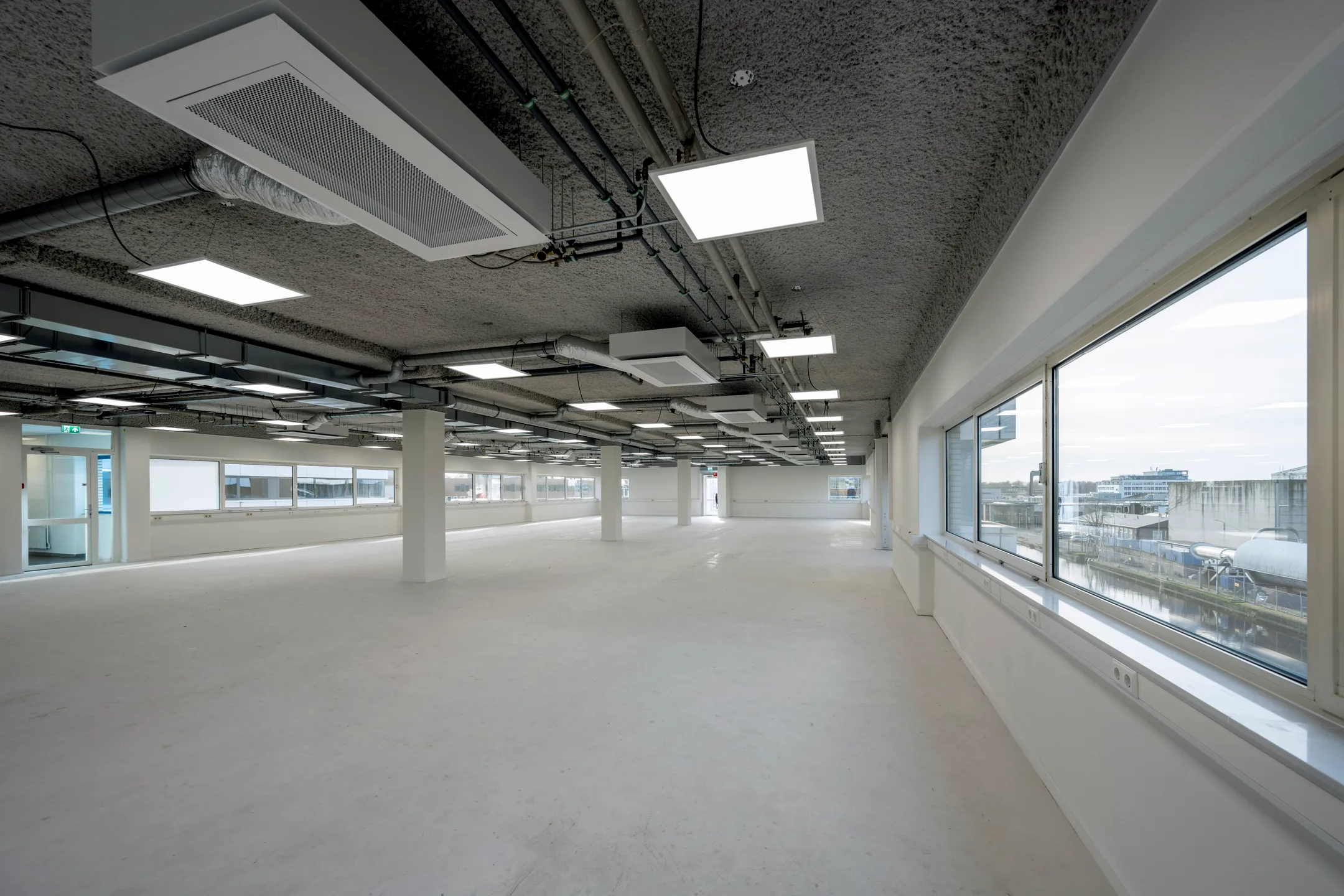 Leeg kantoorinterieur met grote ramen en zichtbare plafondinstallaties.