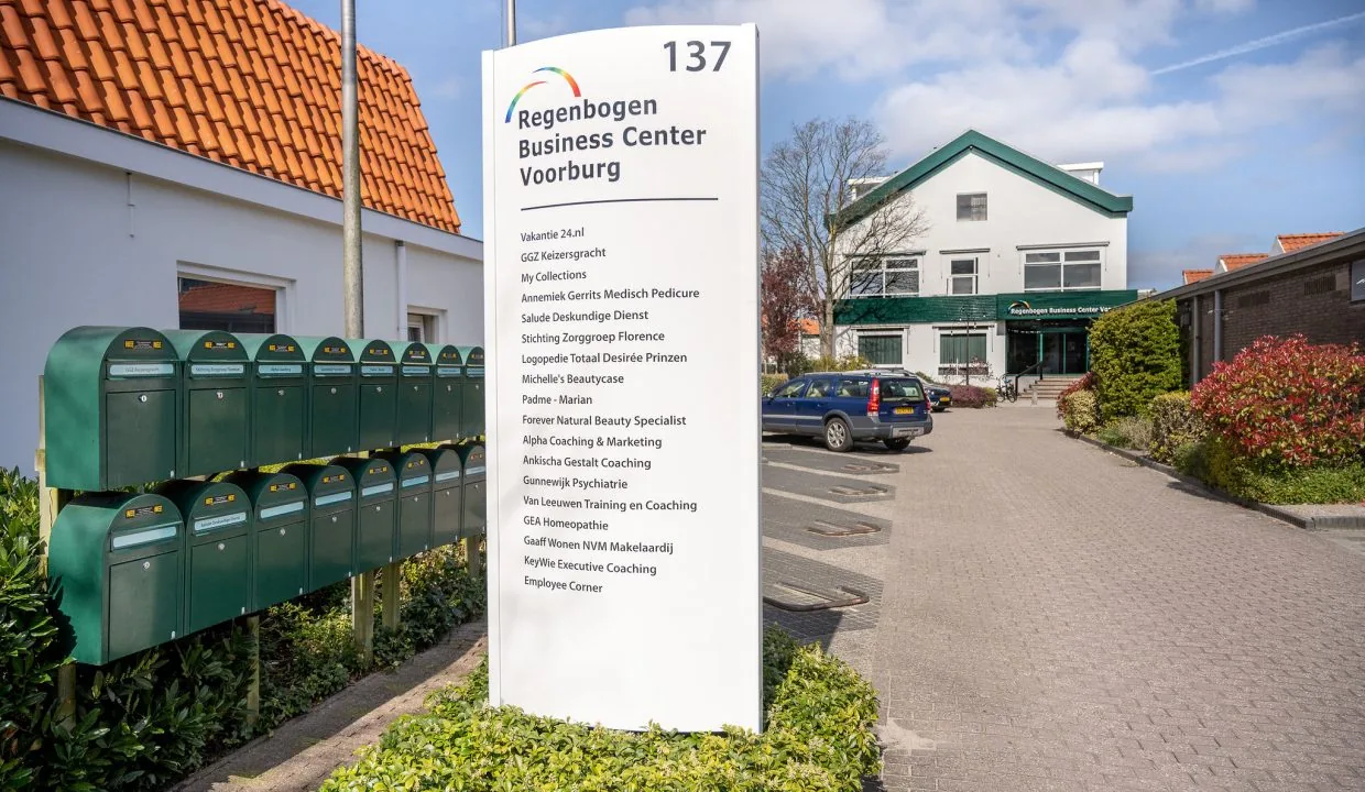 Sign at the entrance of Regenbogen Business Center Voorburg displaying company names, with mailboxes and a parking lot in front of the building.