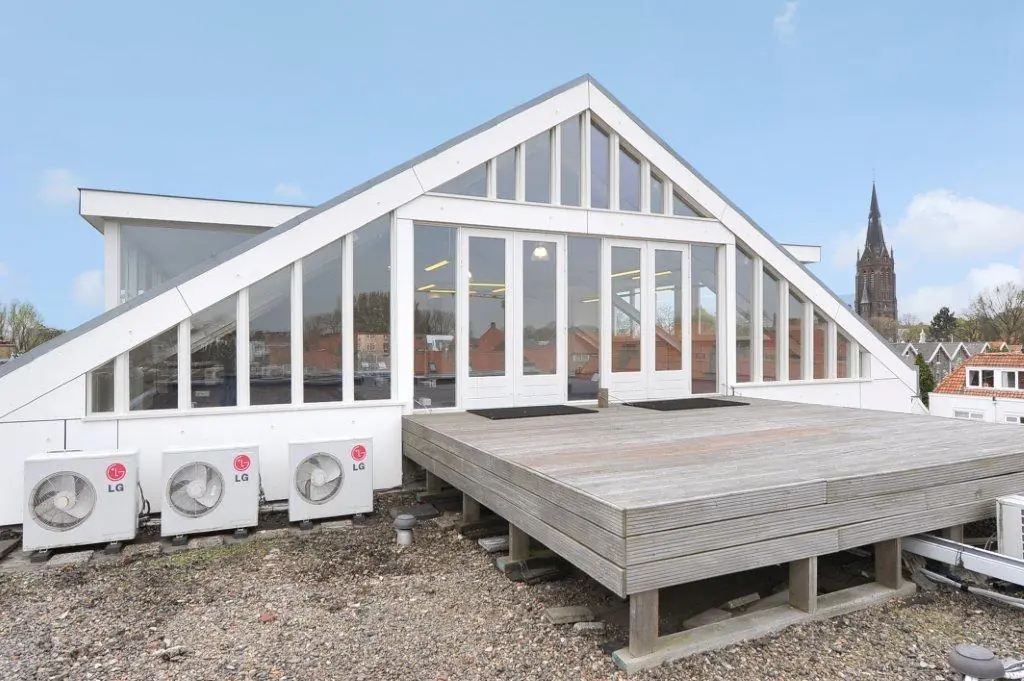 Modern rooftop structure with triangular glass facade and wooden terrace at Oosteinde, featuring air conditioning units and a church tower in the background.