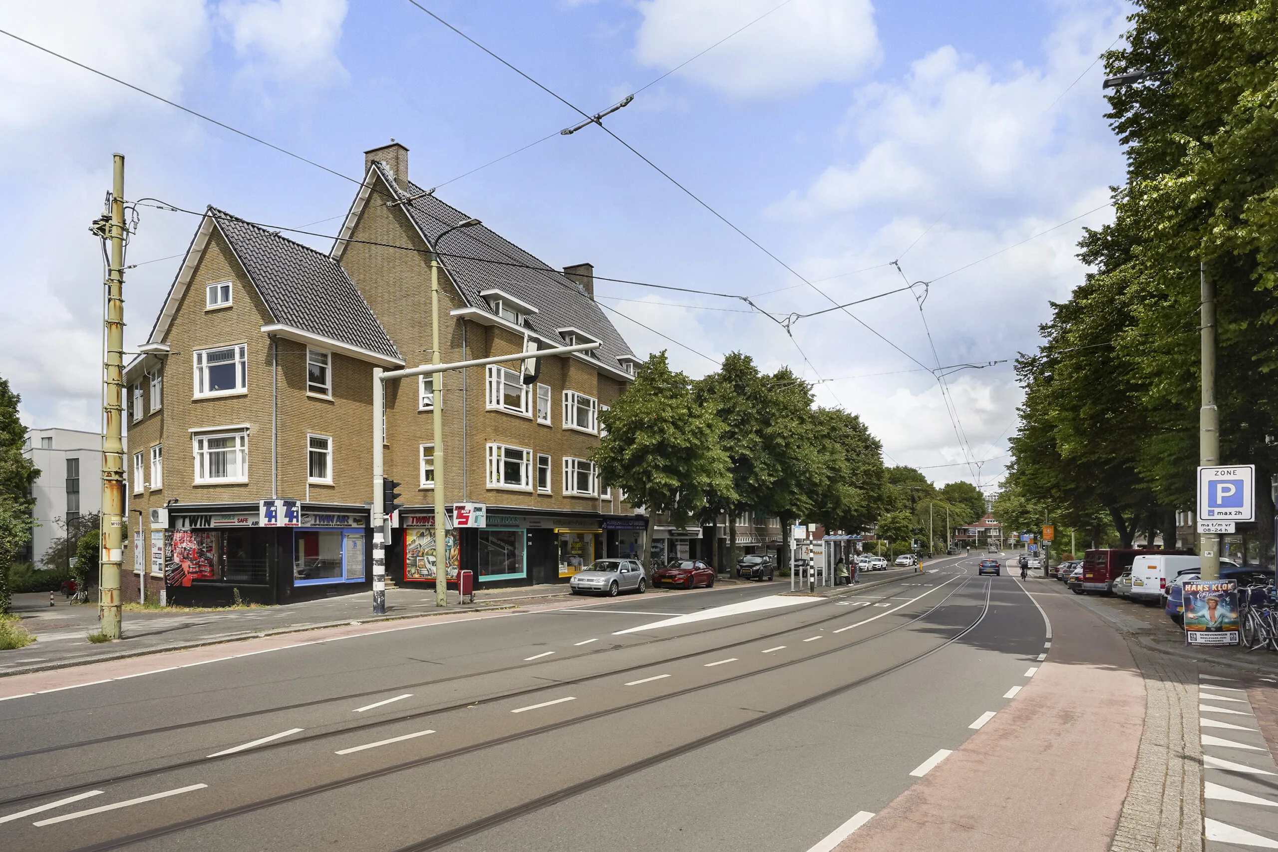 Straatbeeld van de Prinses Mariannelaan met winkels, geparkeerde auto's en tramsporen in een groene woonomgeving.
