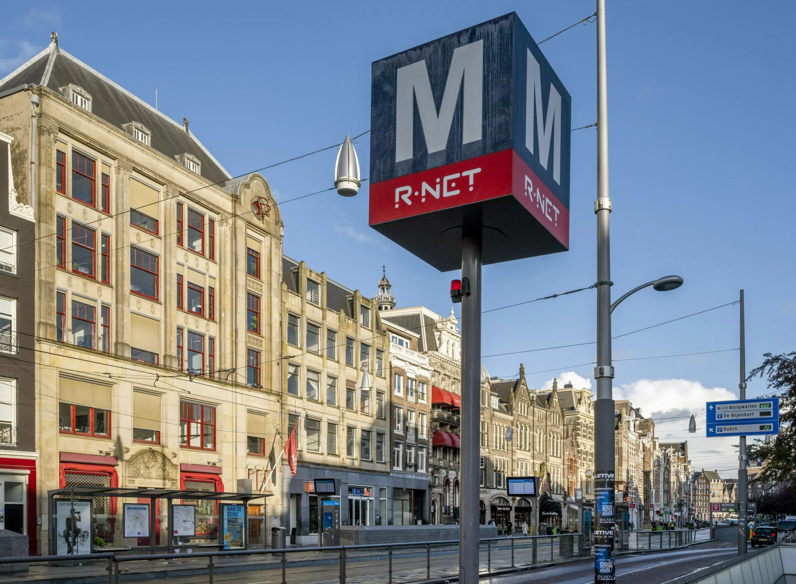 Metro entrance sign for R-NET at Rokin street in Amsterdam with historic buildings in the background.