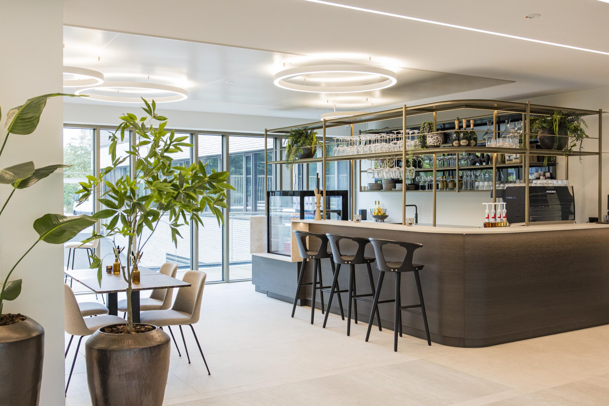 Modern lounge area with a stylish bar, high chairs, indoor plants, and large windows providing natural light.
