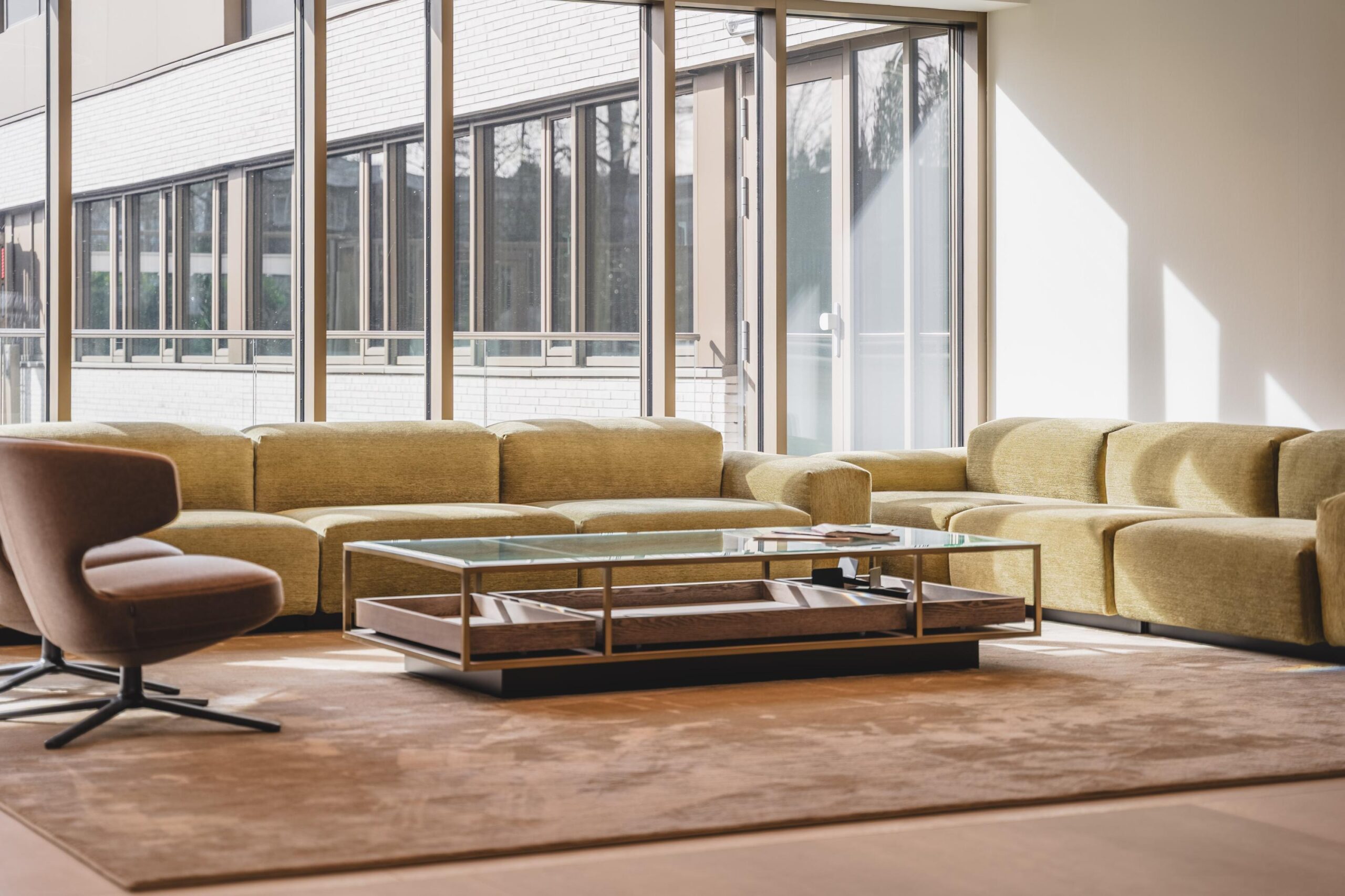 Modern lounge area with green Walborg sectional sofas, a glass coffee table, and a brown armchair in front of large windows.
