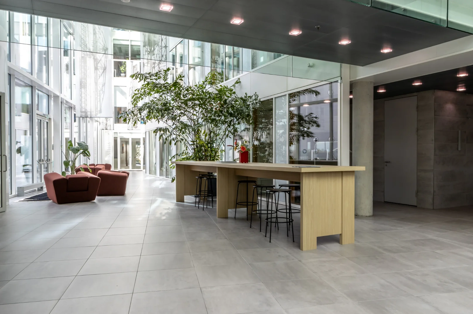 Modern, light-filled office lobby on Pedro de Medinalaan with indoor plants, lounge chairs, and a long wooden table with bar stools.