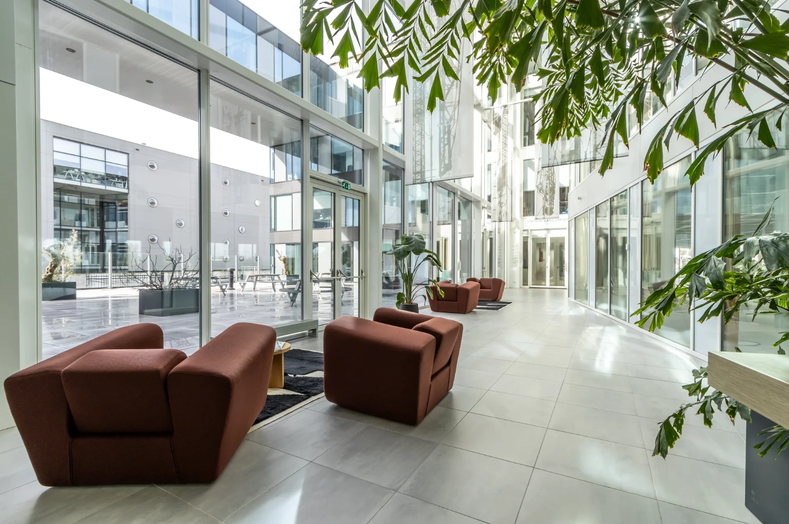 Modern glass atrium with brown lounge chairs, indoor plants, and a view of the Pedro de Medinalaan office courtyard.