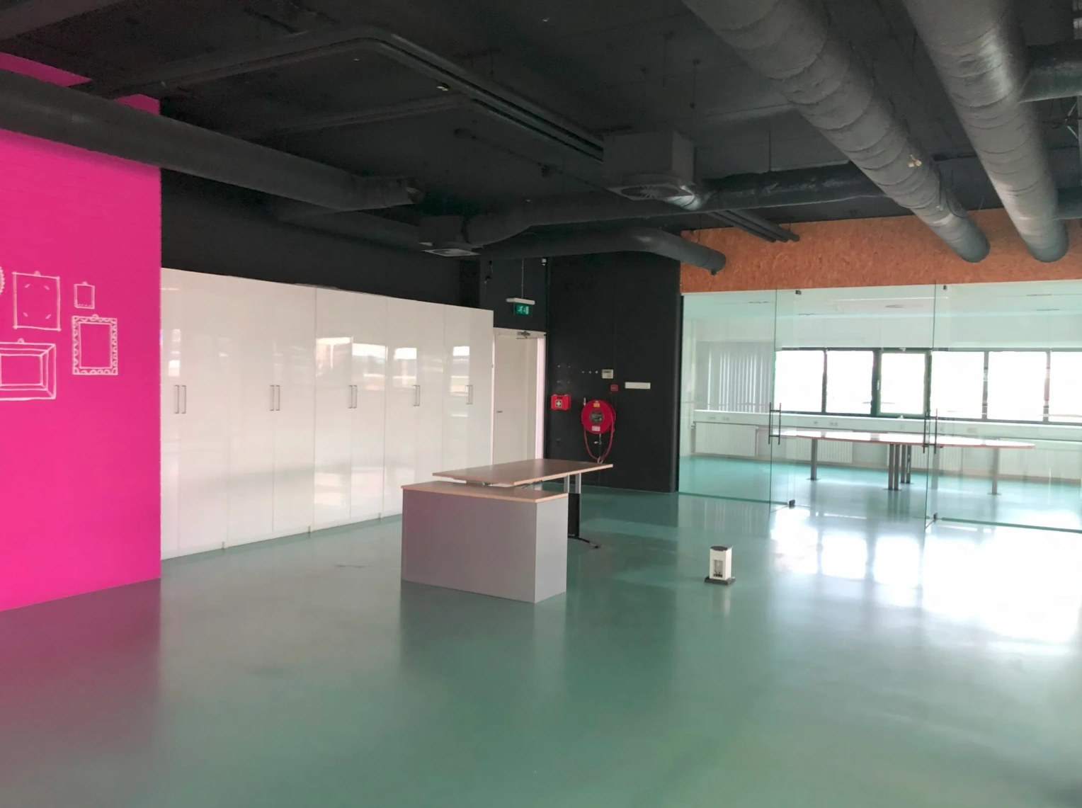 Modern office space on Zekeringstraat with a pink accent wall, white storage cabinets, glass partition, and exposed black ceiling pipes.
