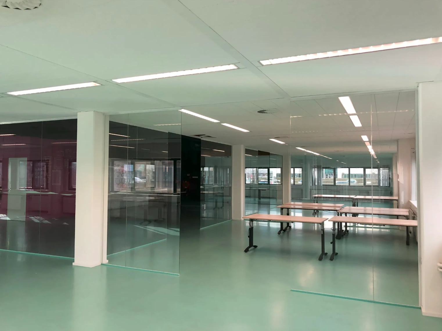 Modern office space on Zekeringstraat with glass walls, fluorescent lighting, and several tables on a mint green floor.