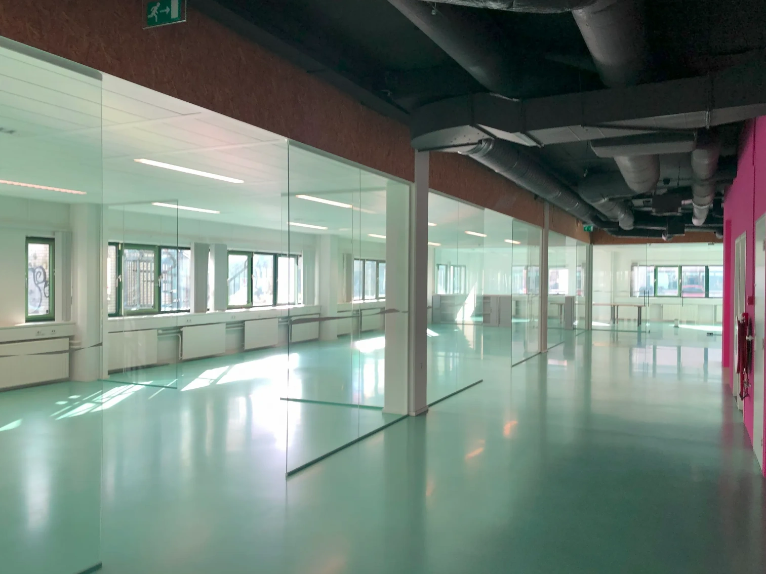 Bright, modern office hallway on Zekeringstraat with glass walls, teal flooring, and exposed ceiling pipes.