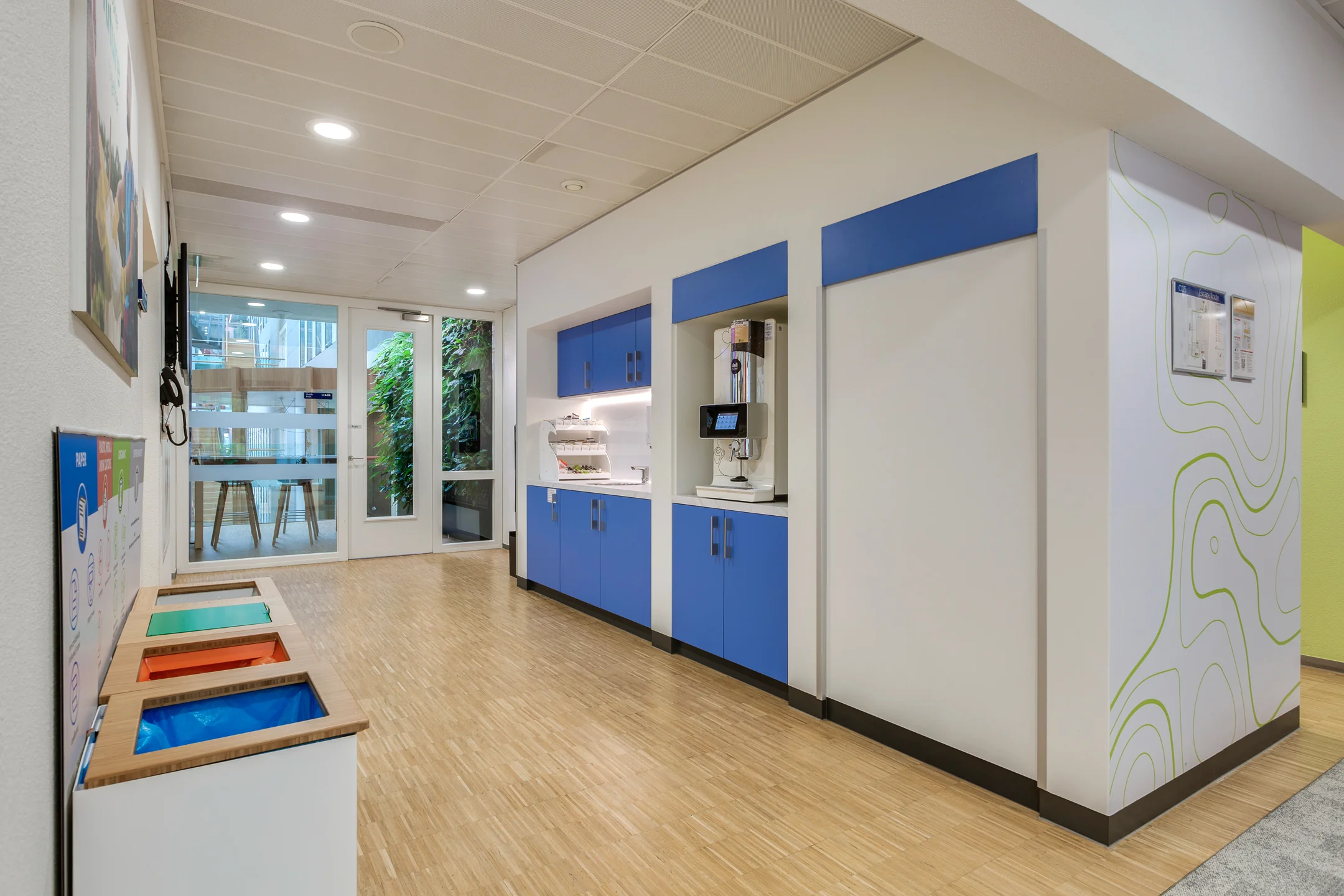 Modern office break area on Carel van Bylandtlaan with blue cabinets, a coffee machine, recycling bins, and glass doors leading to a green courtyard.
