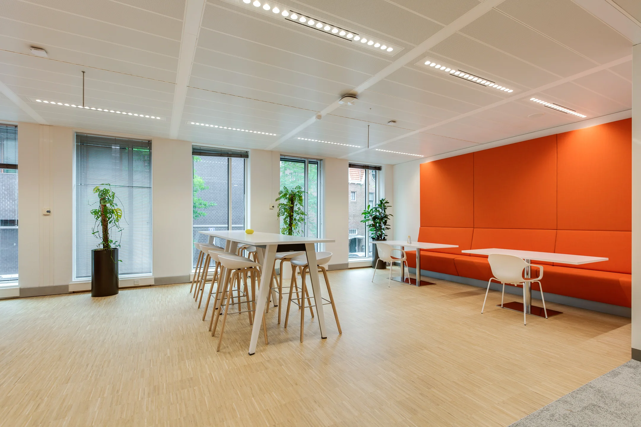 Modern office lounge area on Carel van Bylandtlaan with high tables, orange wall seating, and large windows.