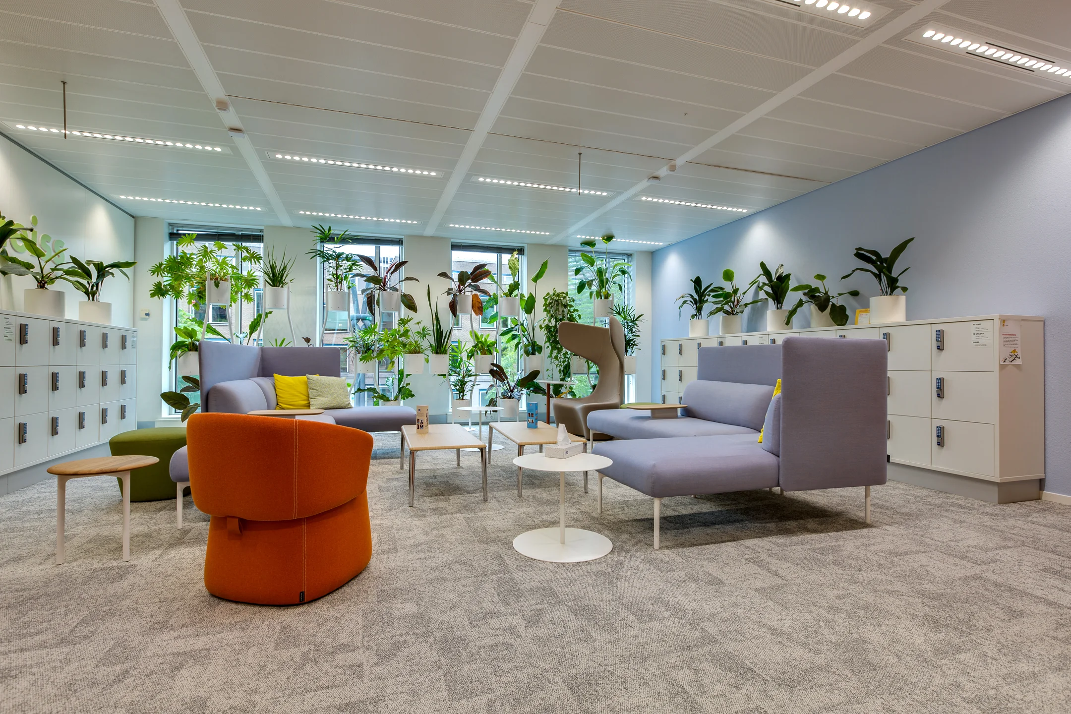 Modern lounge area at Carel van Bylandtlaan with colorful seating, white lockers, and abundant indoor plants.