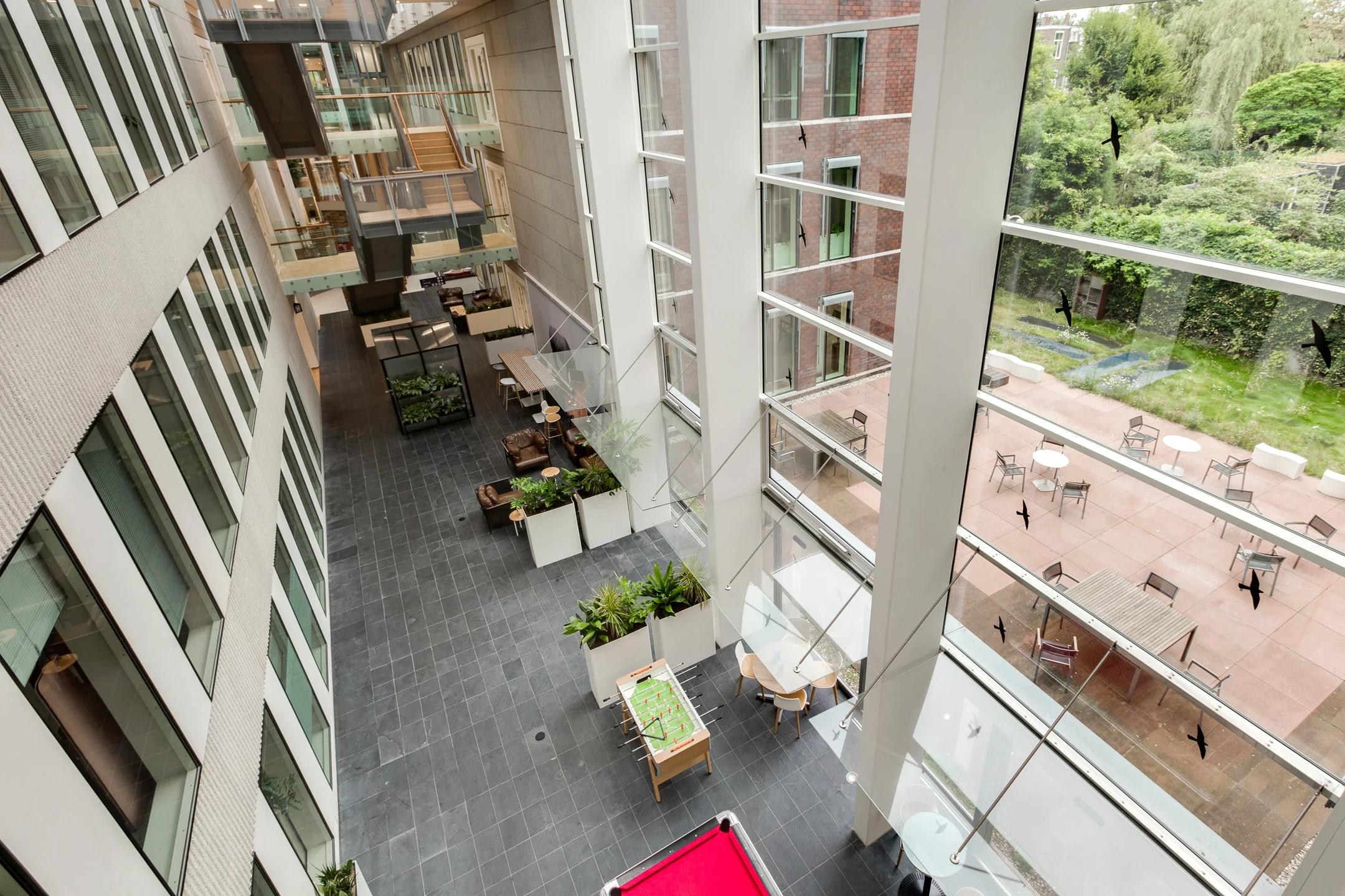Modern atrium interior of a building on Carel van Bylandtlaan with seating areas, greenery, and large windows overlooking an outdoor terrace.