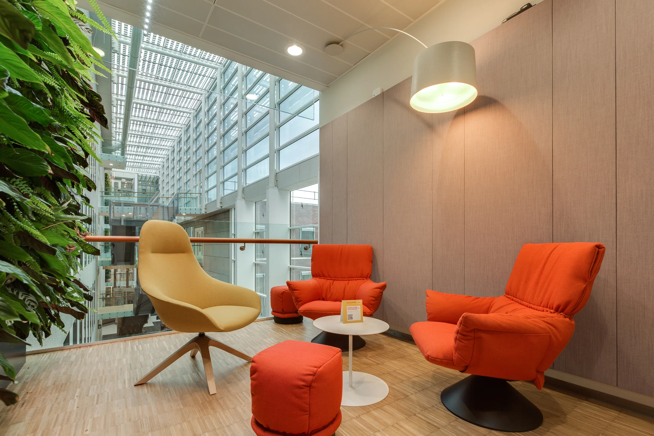 Modern seating area with orange and beige lounge chairs at Carel van Bylandtlaan, in a bright atrium with glass walls and a green plant wall.