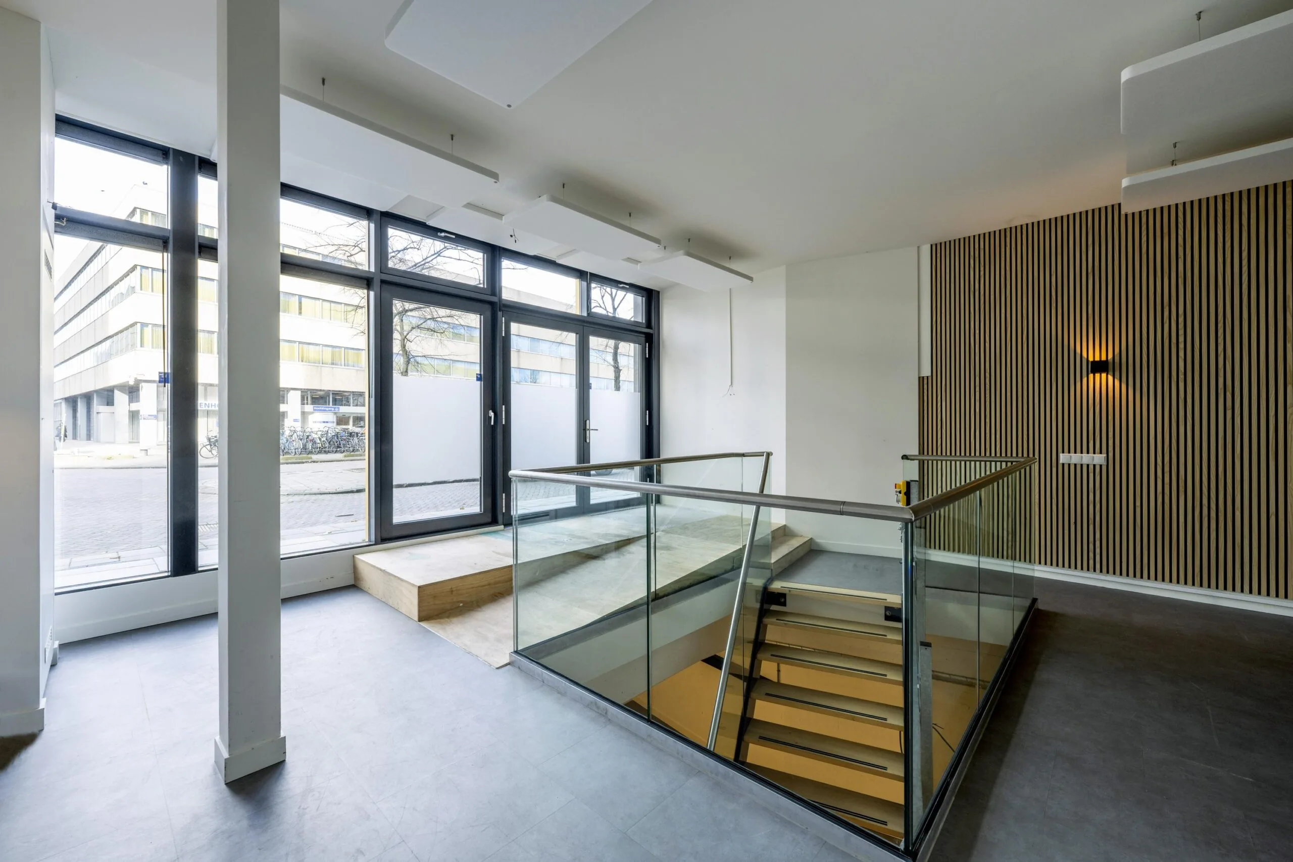 Modern entrance area of a building on James Wattstraat with glass doors, large windows, and a staircase with glass railing.