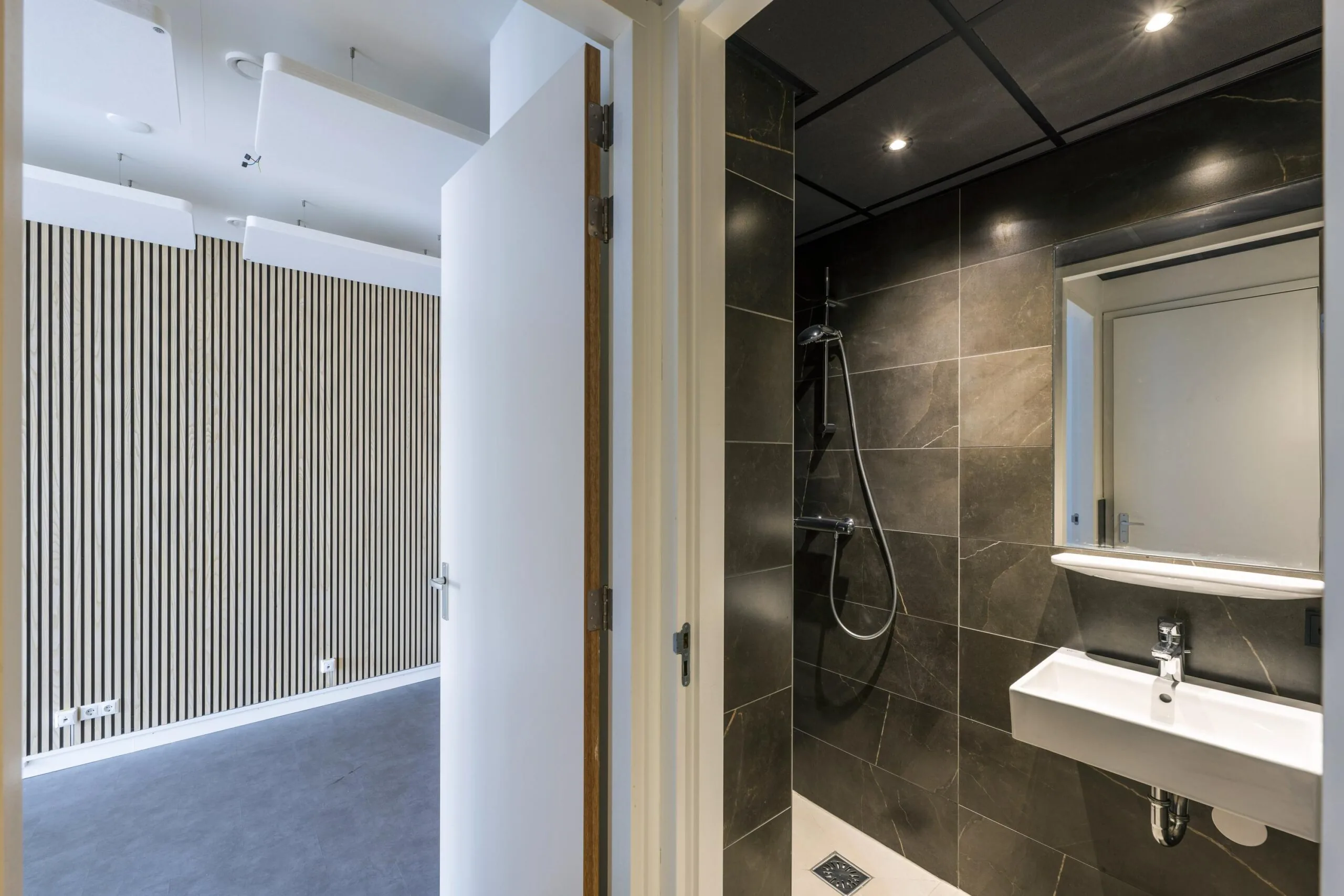 Modern bathroom with dark tiles and walk-in shower next to a minimalistic room in James Wattstraat.