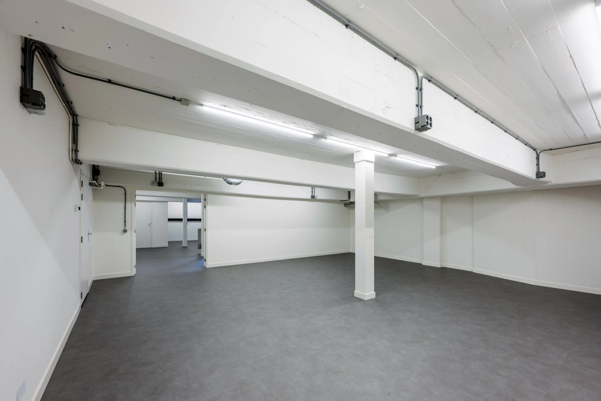 Spacious, empty basement interior at James Wattstraat with white walls, gray flooring, and exposed ceiling pipes.