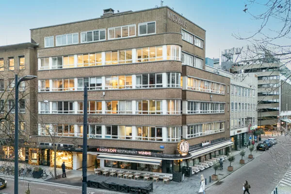 Een hoekig kantoorgebouw genaamd "Minervahuis" met een café-restaurant-wijnbar op de begane grond in een stedelijke omgeving.