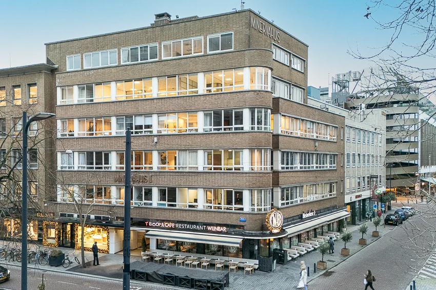 Een hoekig kantoorgebouw genaamd "Minervahuis" met een café-restaurant-wijnbar op de begane grond in een stedelijke omgeving.