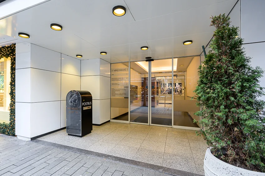 Ingang van een modern gebouw met glazen deuren en een postbus, naast een versierde plantenbak.