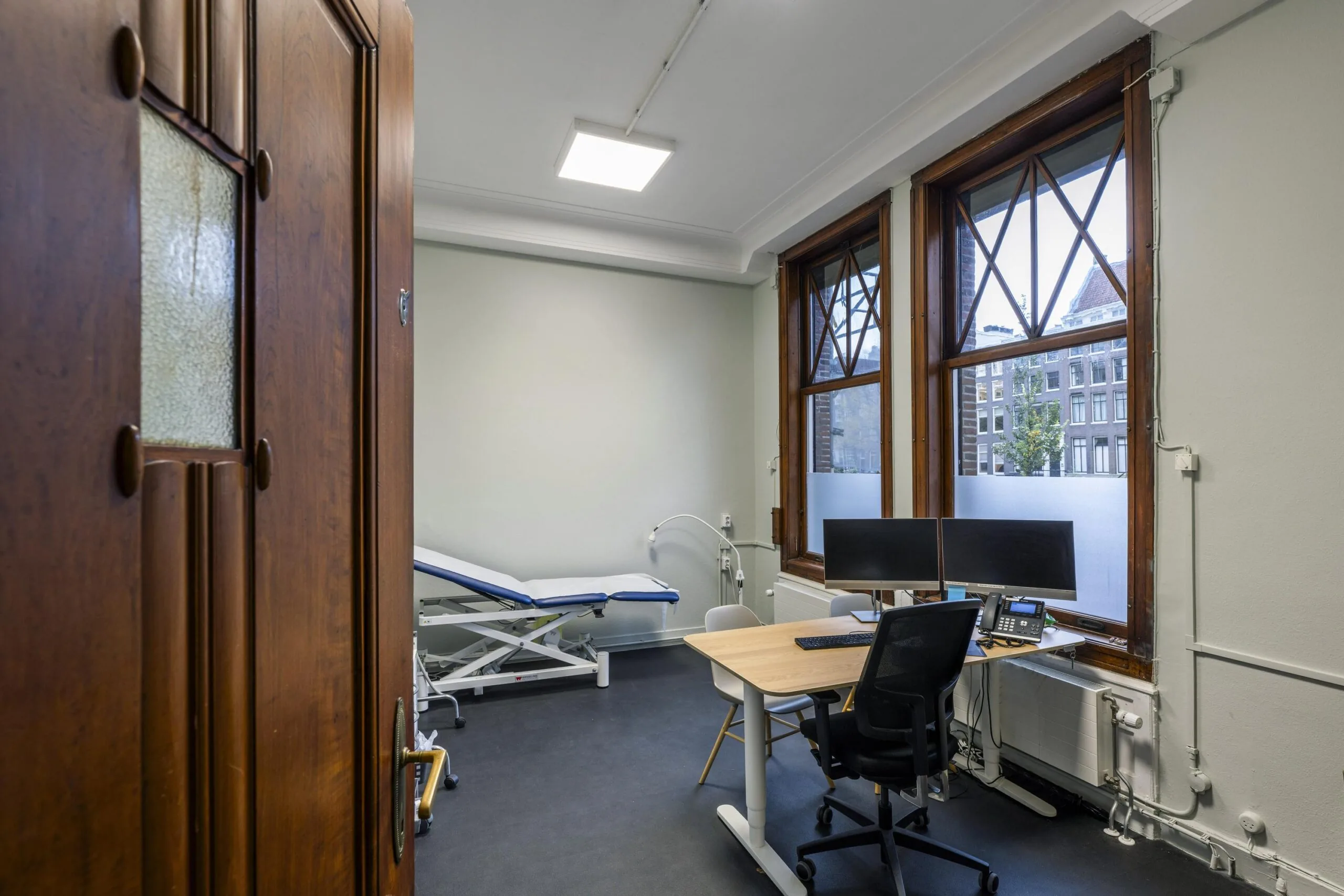 Modern medical consultation room at Westermarkt with a desk, dual monitors, and an examination table.