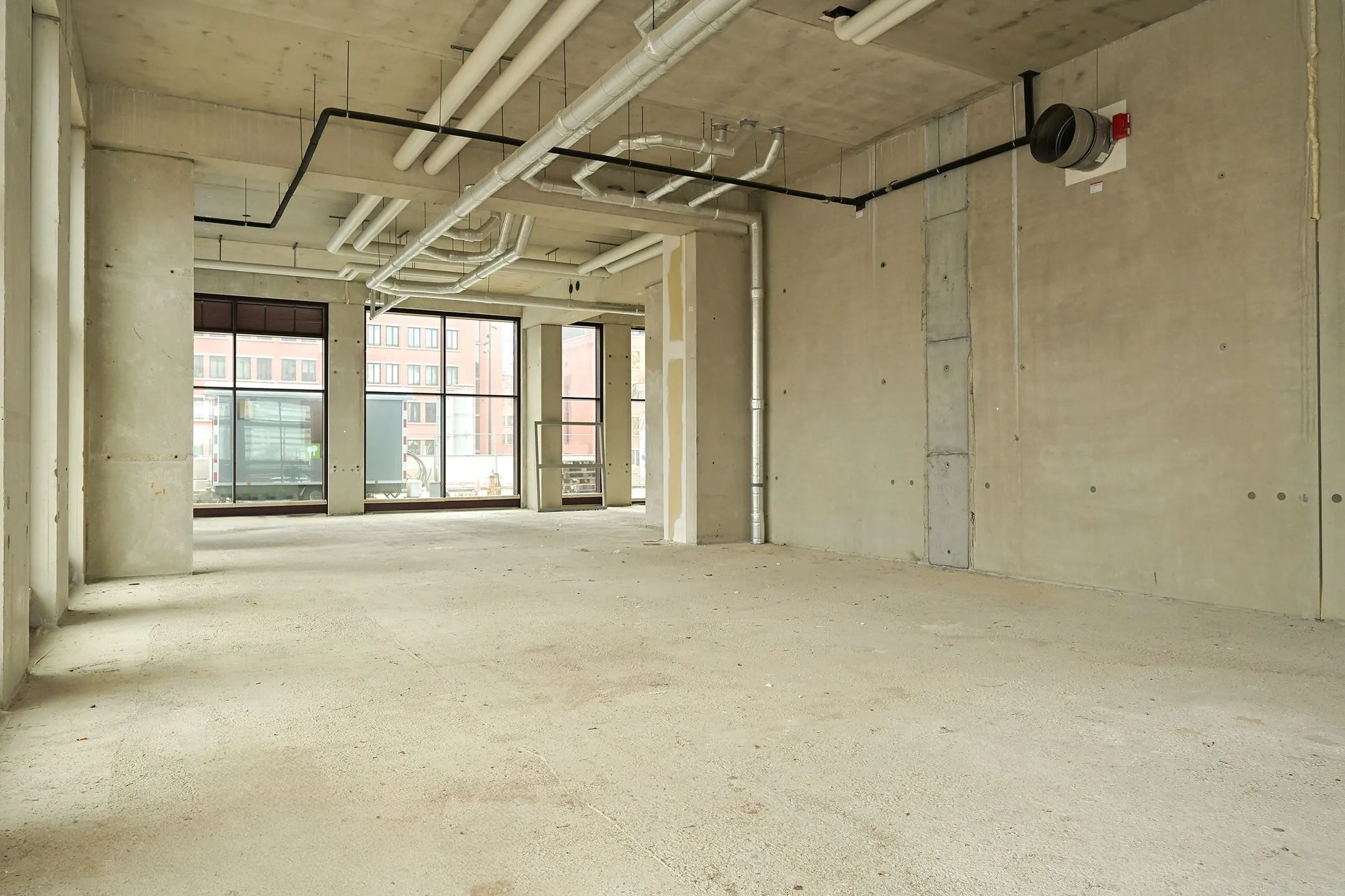 Empty commercial space on Melkwegstraat with exposed concrete walls, large windows, and visible ceiling pipes.