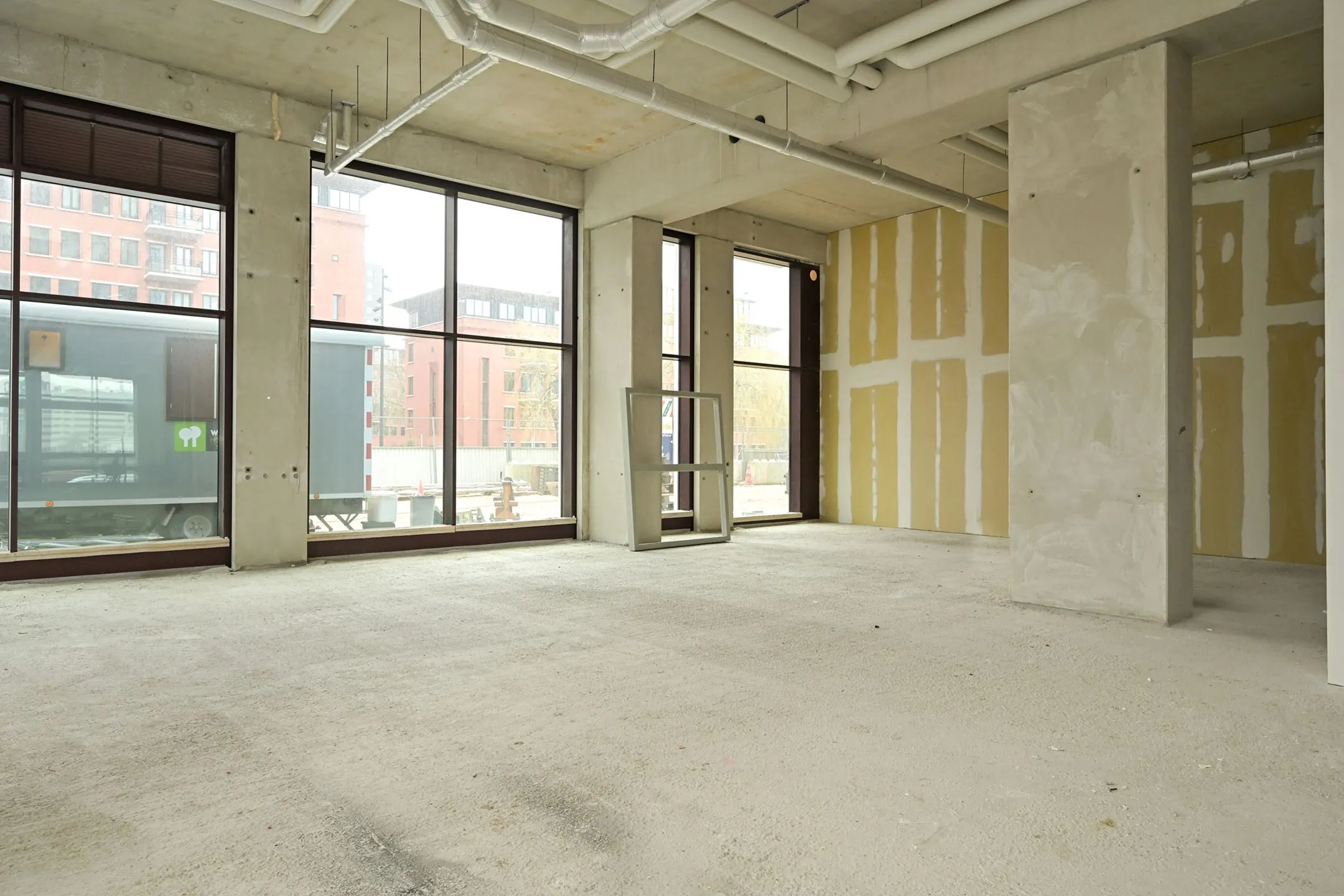 Empty commercial space on Melkwegstraat with unfinished walls, concrete floor, and large windows facing a modern urban street.