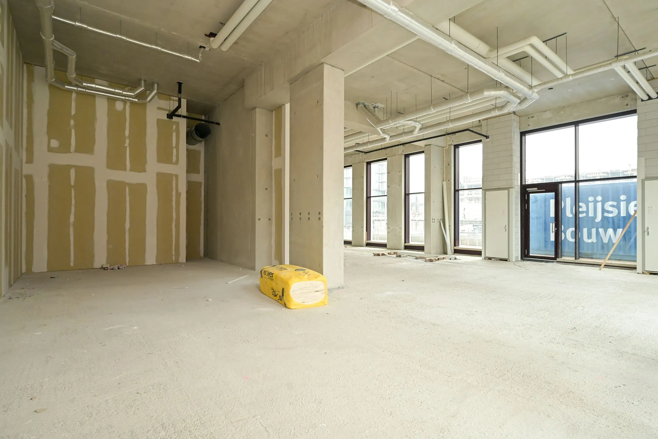 Interior view of an unfinished commercial space on Melkwegstraat with exposed pipes, concrete walls, and large windows.