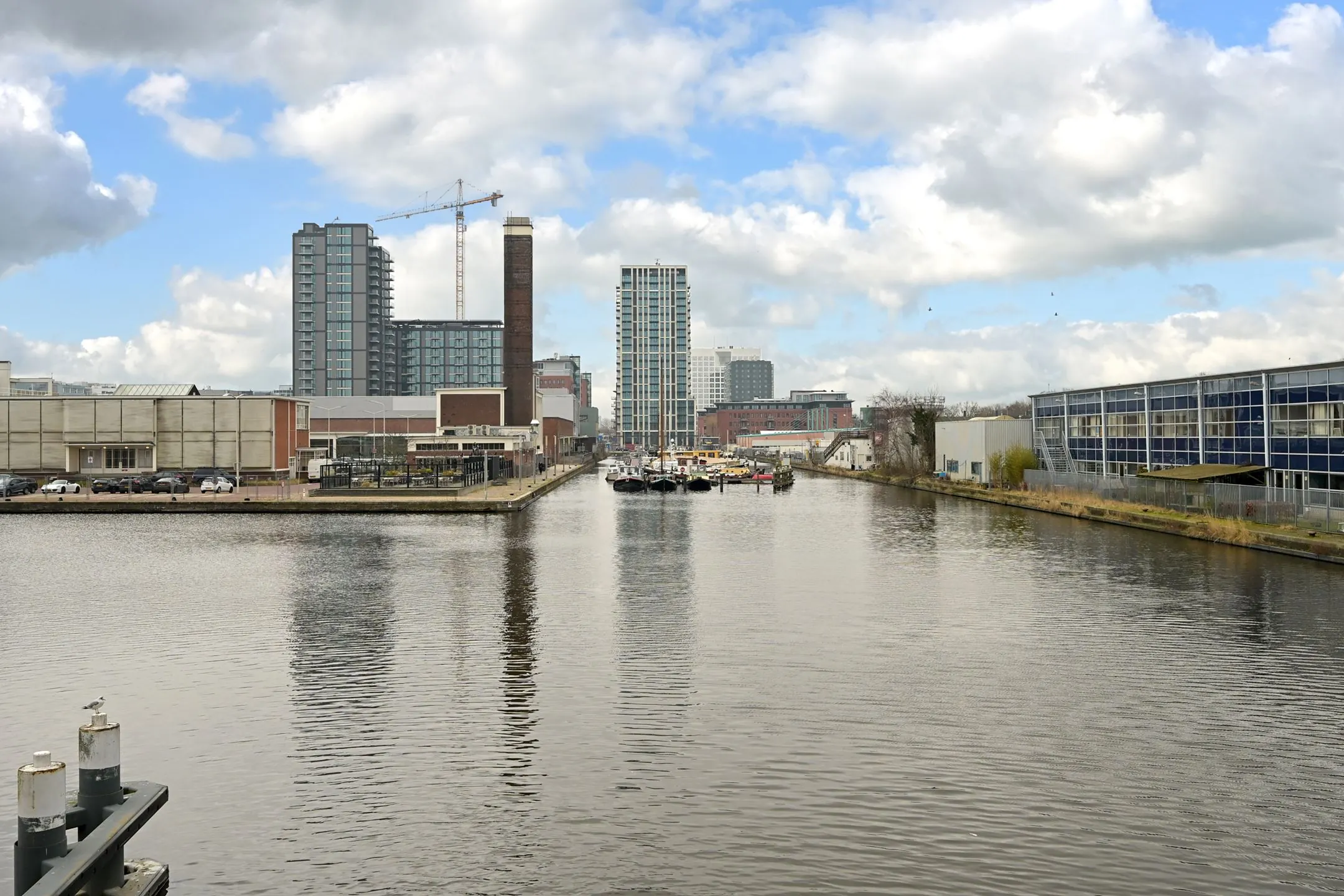 View of the canal at Melkwegstraat with modern high-rise buildings, industrial warehouses, and moored boats under a partly cloudy sky.