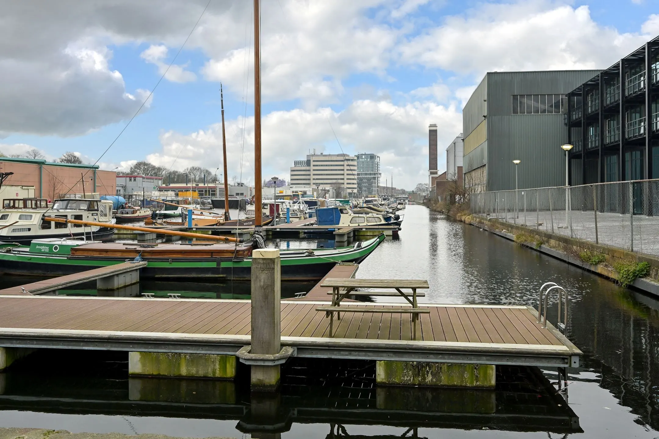 View of boats moored along the canal at Melkwegstraat, with industrial buildings and offices in the background.