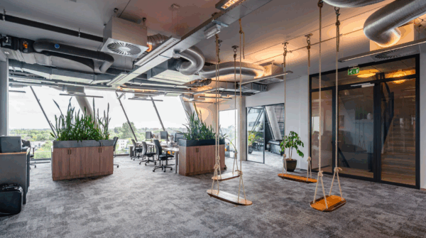 Modern open-plan office space on the Vasumweg with hanging swings, large ceiling pipes, indoor plants, and workstations near large windows.