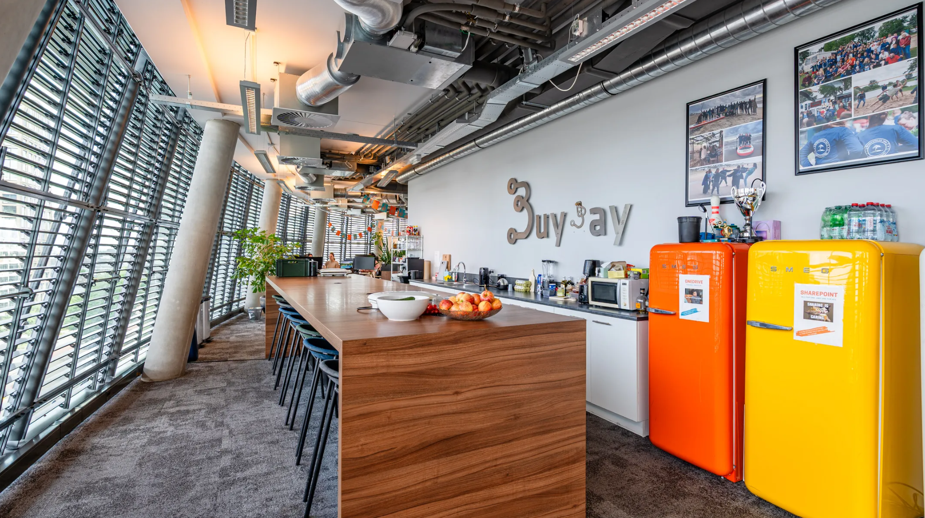 Modern office kitchen area at BuyBay with a long wooden table, colorful SMEG fridges, and industrial-style design.