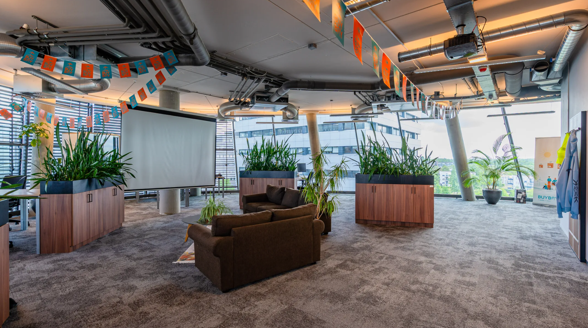 Modern office lounge area at TT Vasumweg with indoor plants, colorful bunting, a projector screen, and large windows offering a city view.