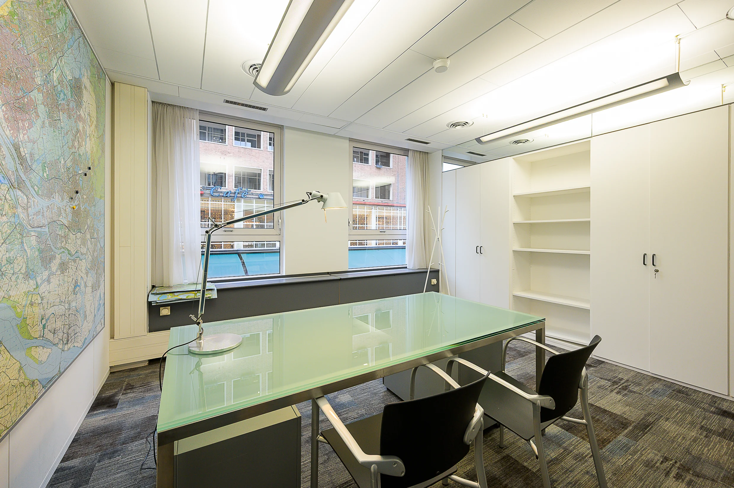 Een modern kantoor met een glazen bureau, stoelen, een wereldkaart aan de muur en uitzicht op een café door het raam.
