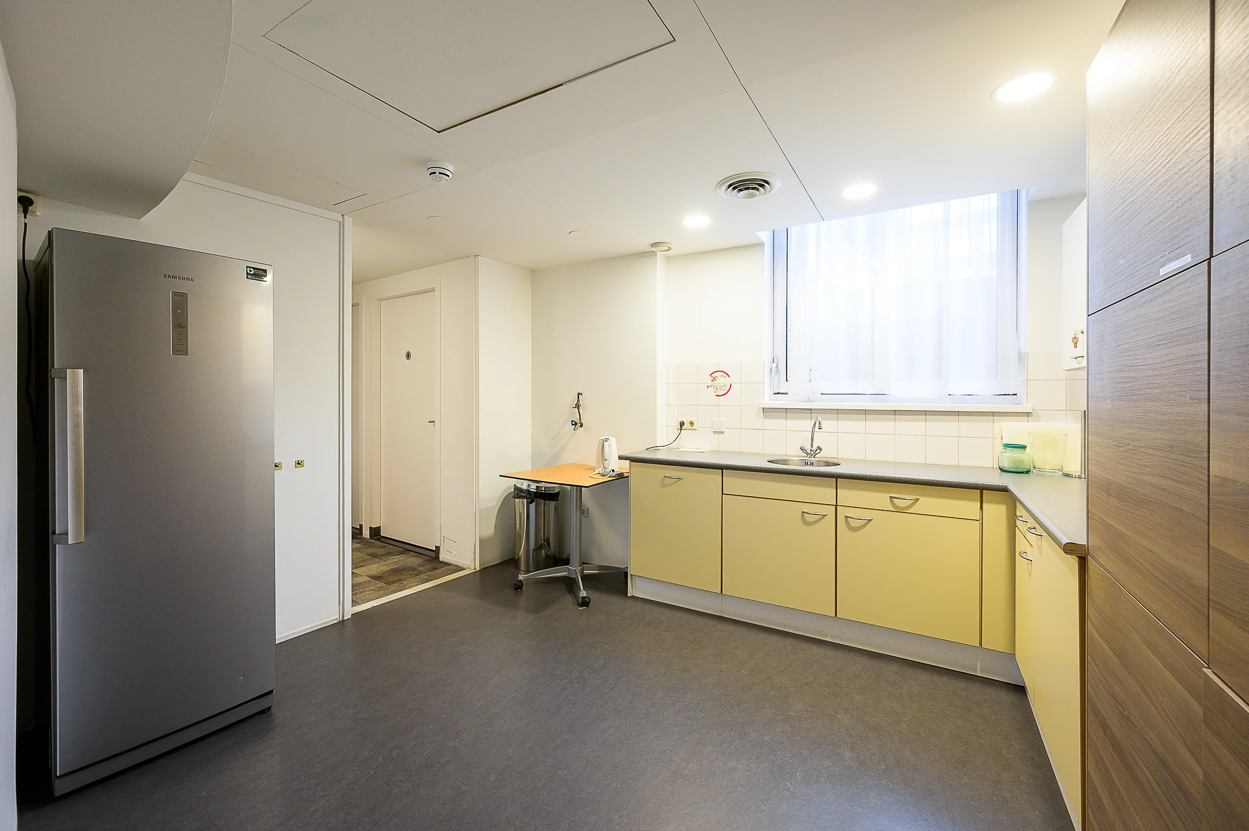 Een moderne keuken met een koelkast, wastafel, kastjes en een klein tafeltje onder een raam.