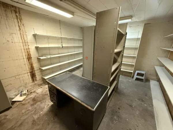 A small, worn storage room on Katendrechtse Lagedijk with empty shelves, a metal desk, and stained concrete walls.