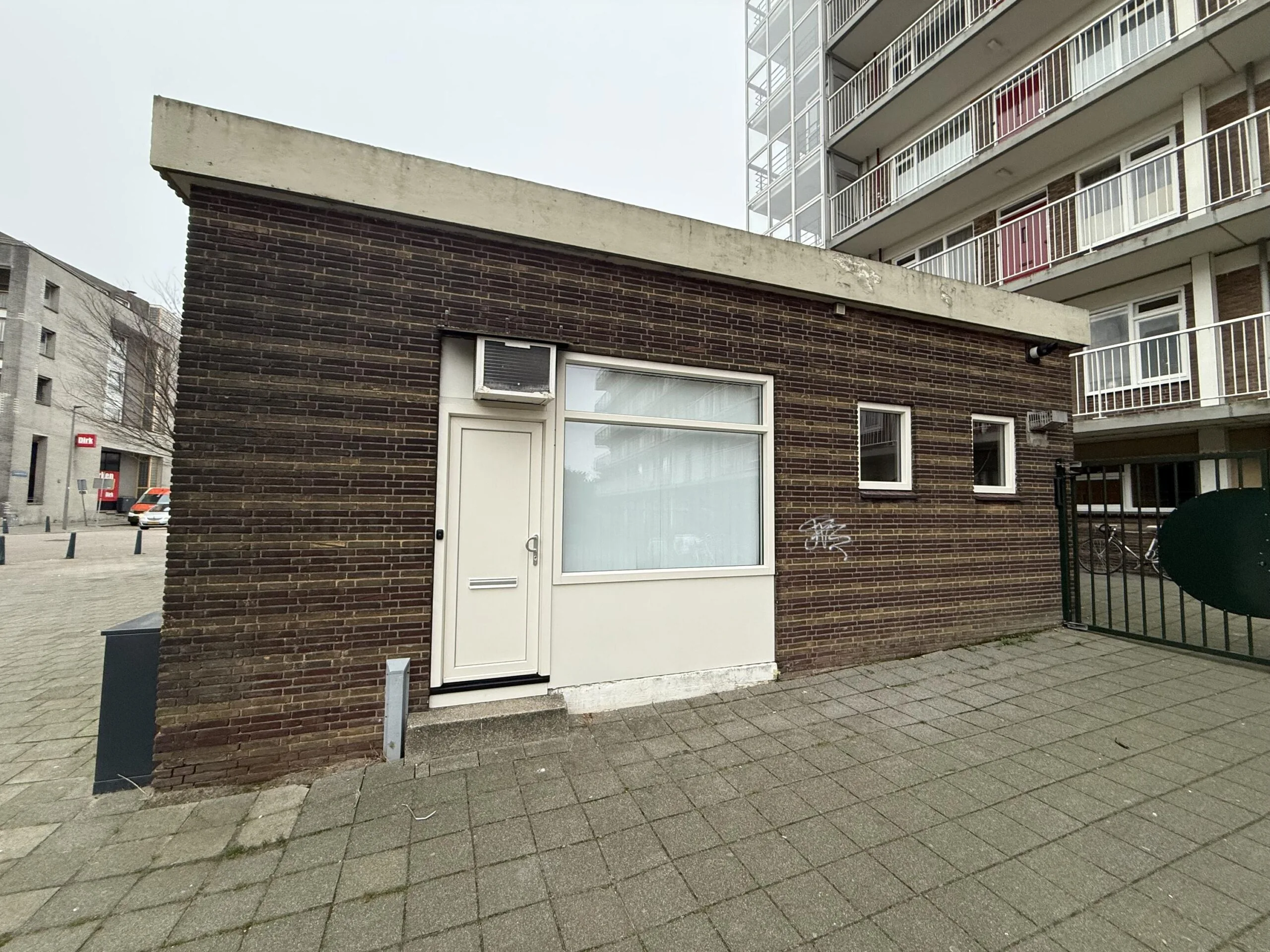 Small brick building with a white door and window on Katendrechtse Lagedijk, next to a residential apartment block.