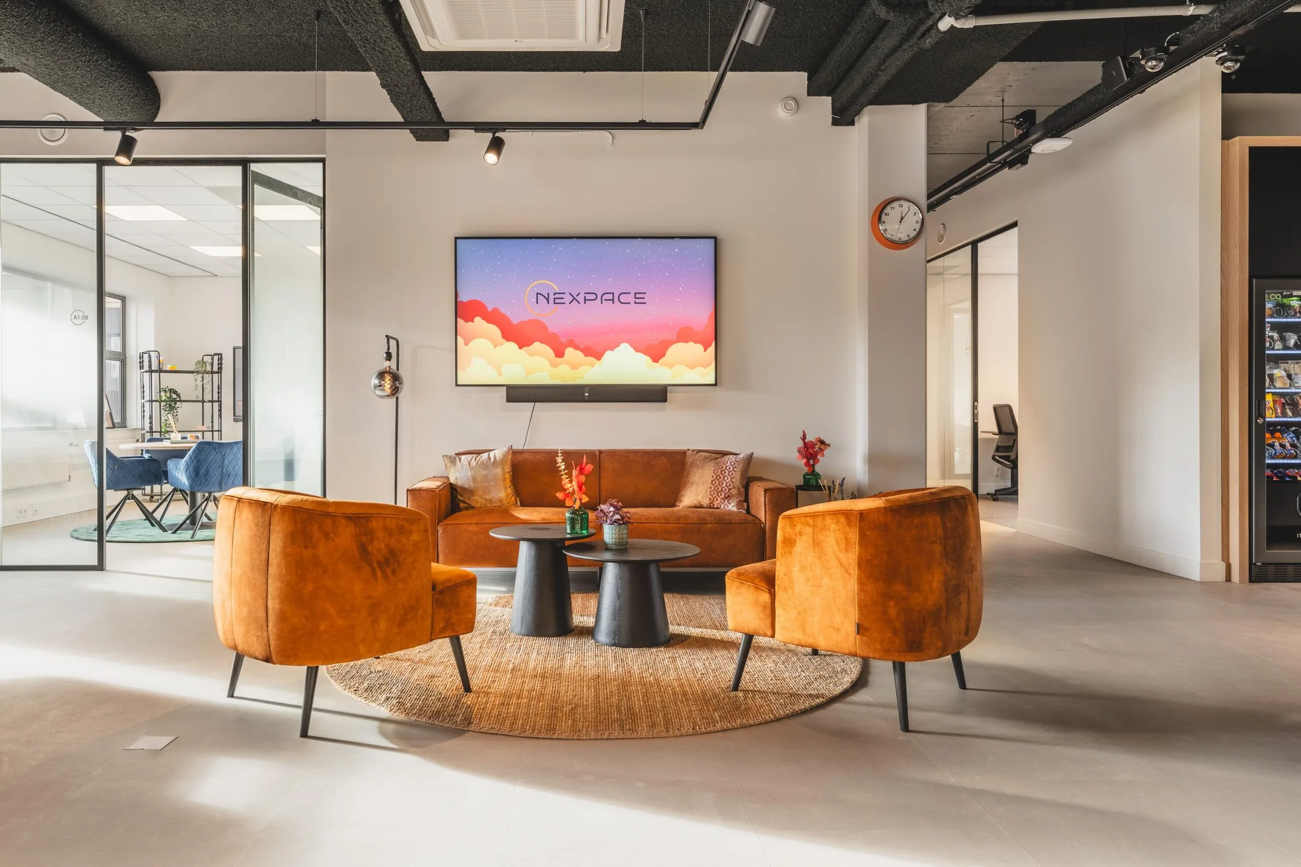 Modern office lounge area at Paasheuvelweg with orange velvet chairs, a round rug, and a Nexpace screen on the wall.