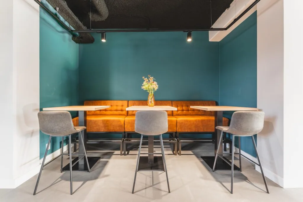 Modern seating area at Paasheuvelweg with orange upholstered bench, high tables, and gray bar chairs against teal walls.
