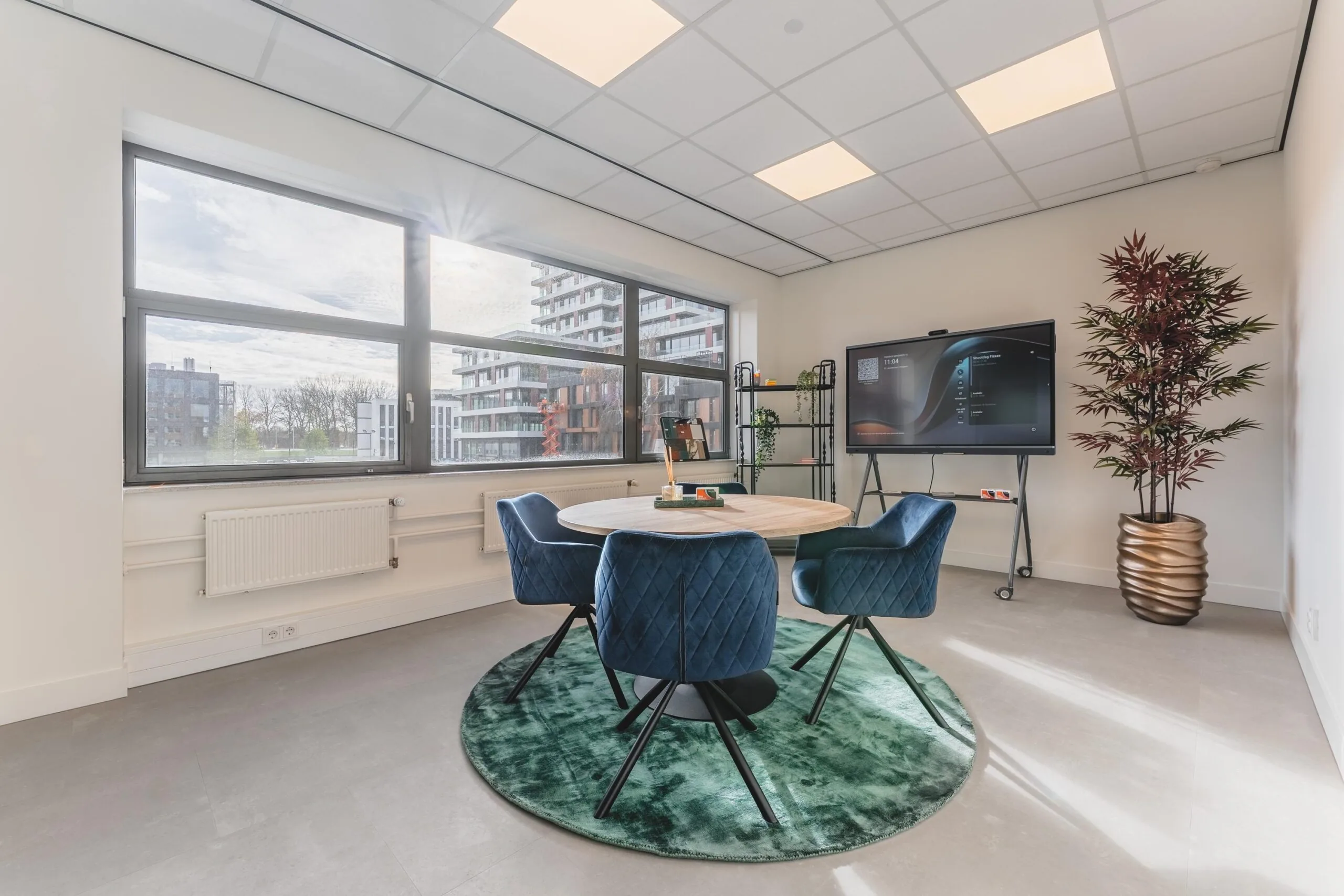 Modern meeting room at Paasheuvelweg with a round table, blue chairs, large window, and digital presentation screen.