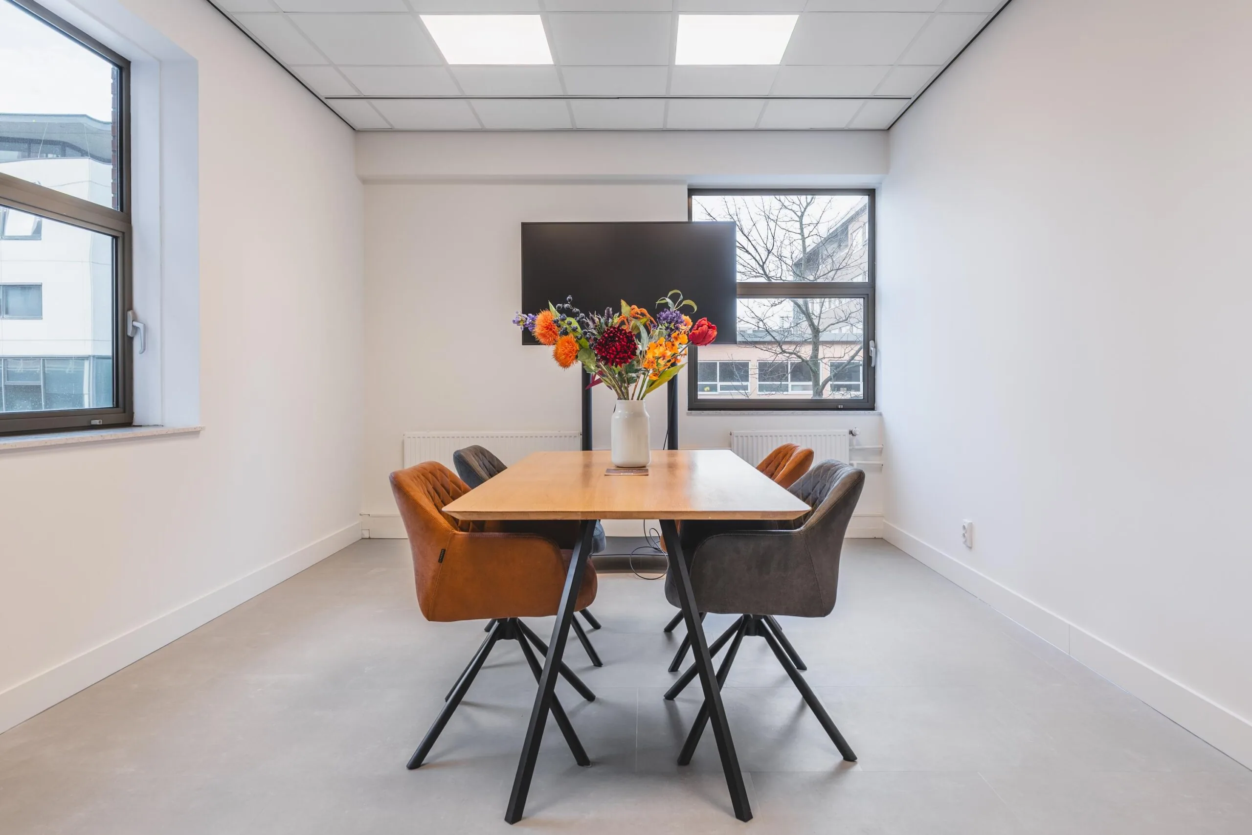 Modern meeting room on Paasheuvelweg with a wooden table, colorful chairs, a large screen, and a vase of vibrant flowers.