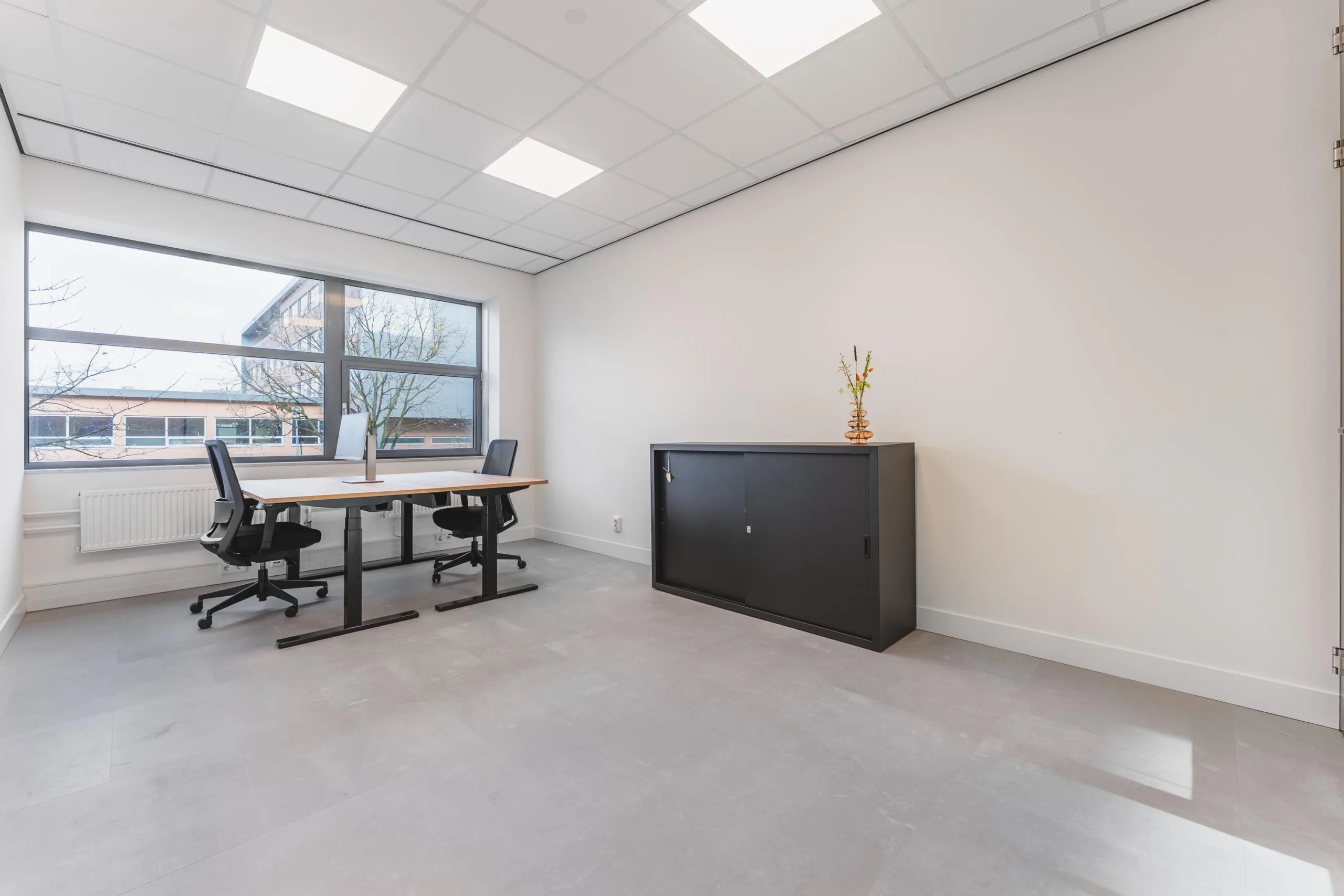 Modern office space at Paasheuvelweg with two desks, ergonomic chairs, large window, and a black cabinet with a decorative vase.