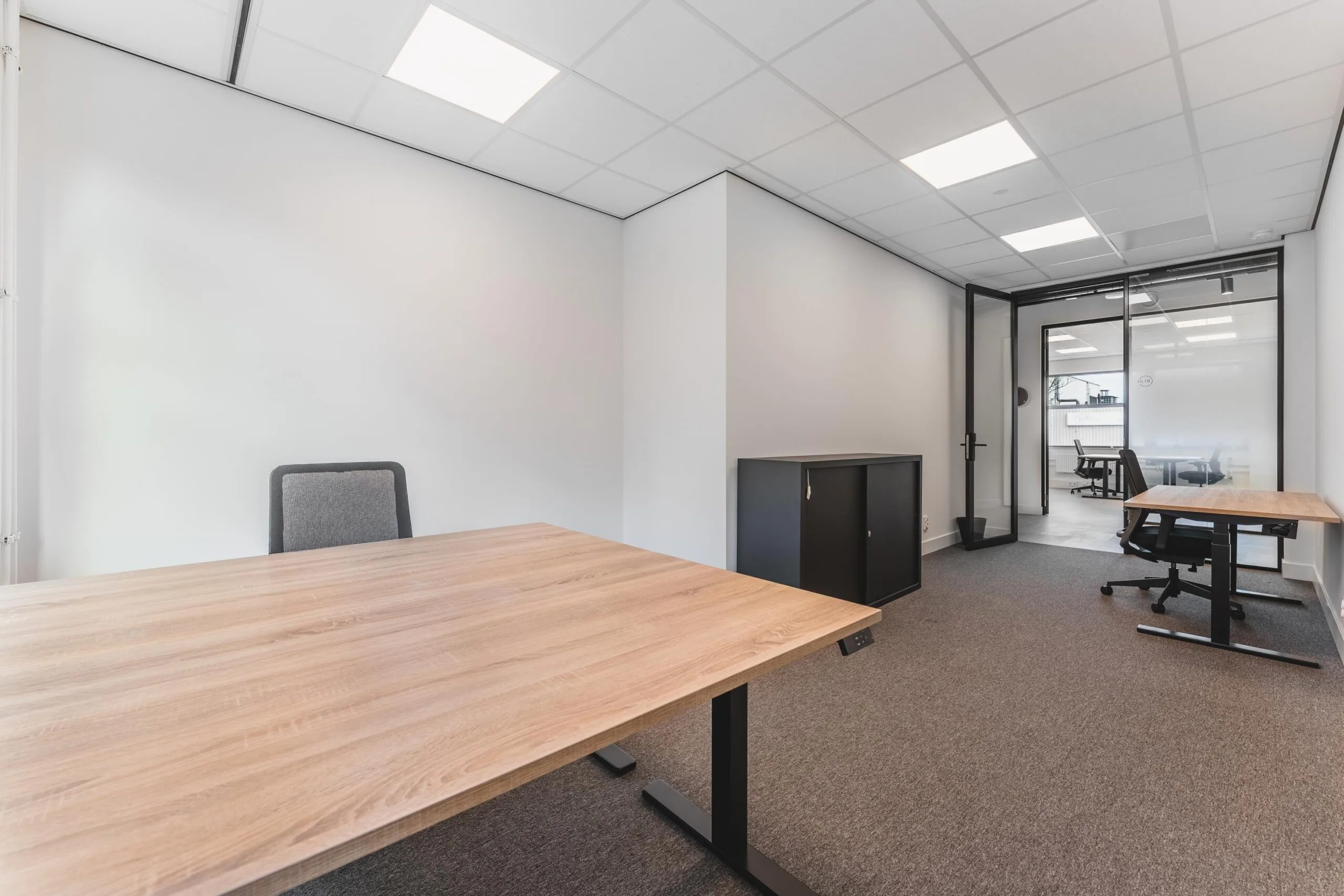 Modern office space at Paasheuvelweg with wooden desks, carpeted floor, and glass partition wall.