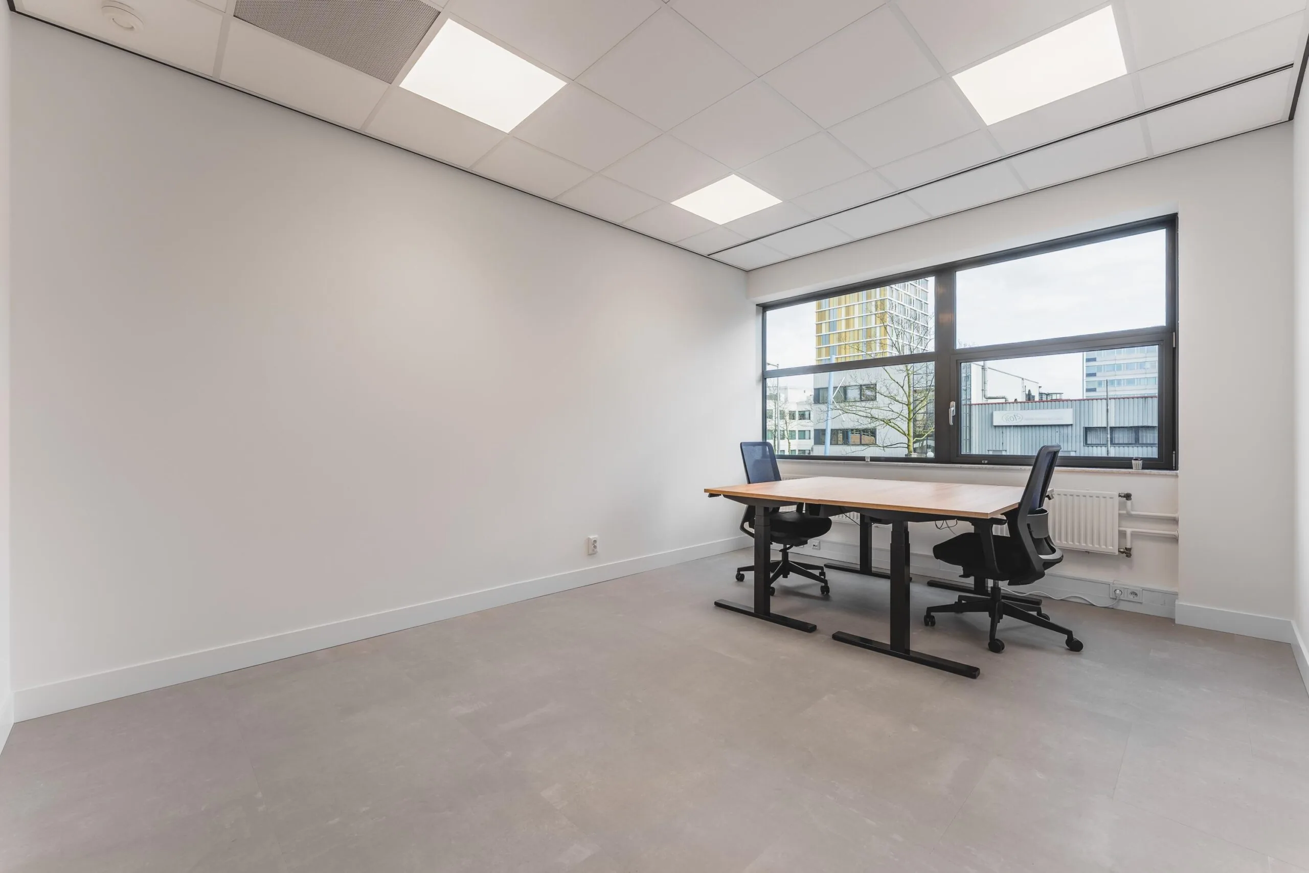 Modern, minimal office space at Paasheuvelweg with two desks and large windows overlooking nearby buildings.