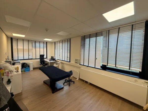 Modern medical examination room at Veteranenlaan with a treatment table, desks, and large windows with blinds.
