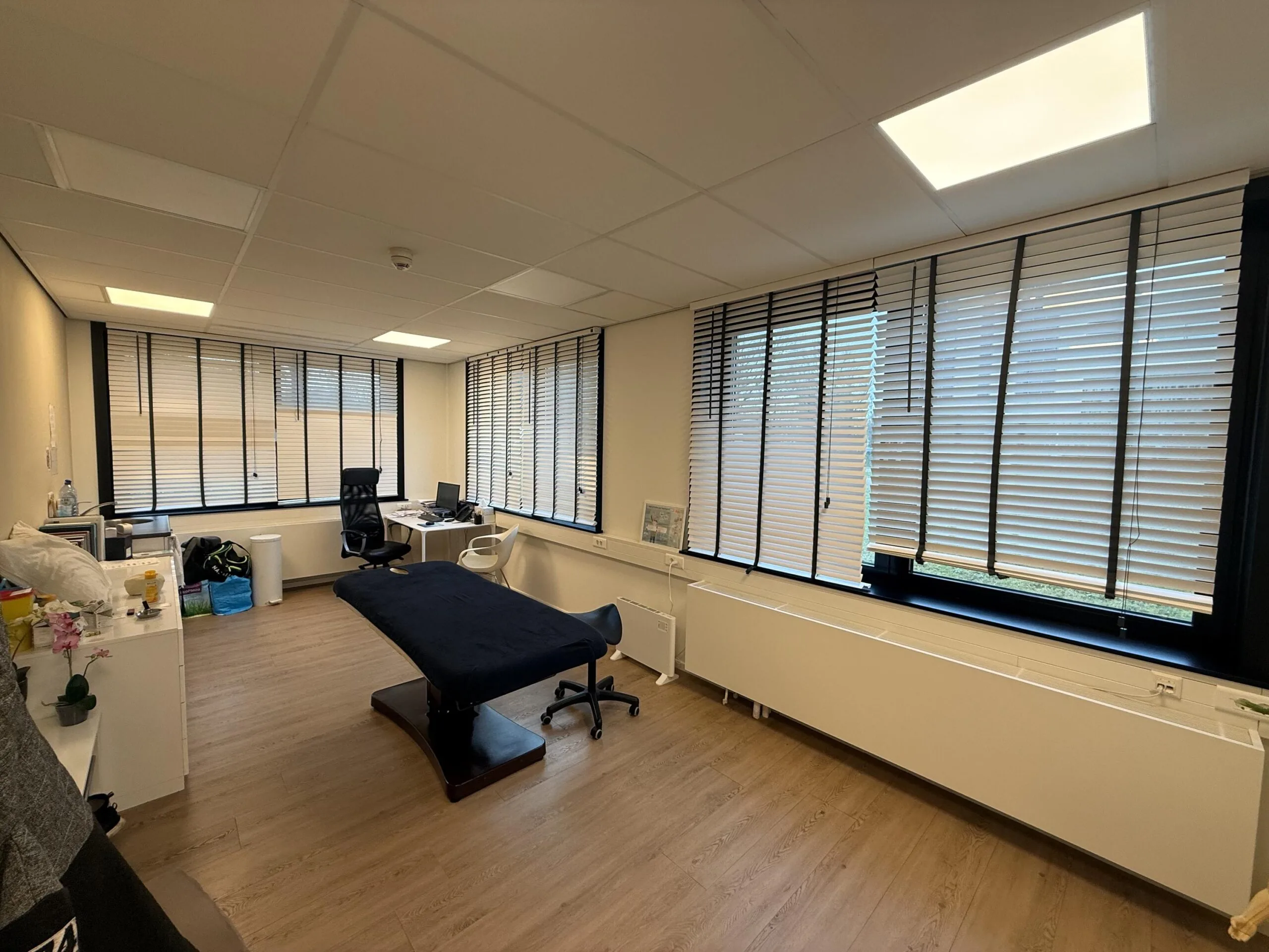 Modern medical examination room at Veteranenlaan with a treatment table, desks, and large windows with blinds.