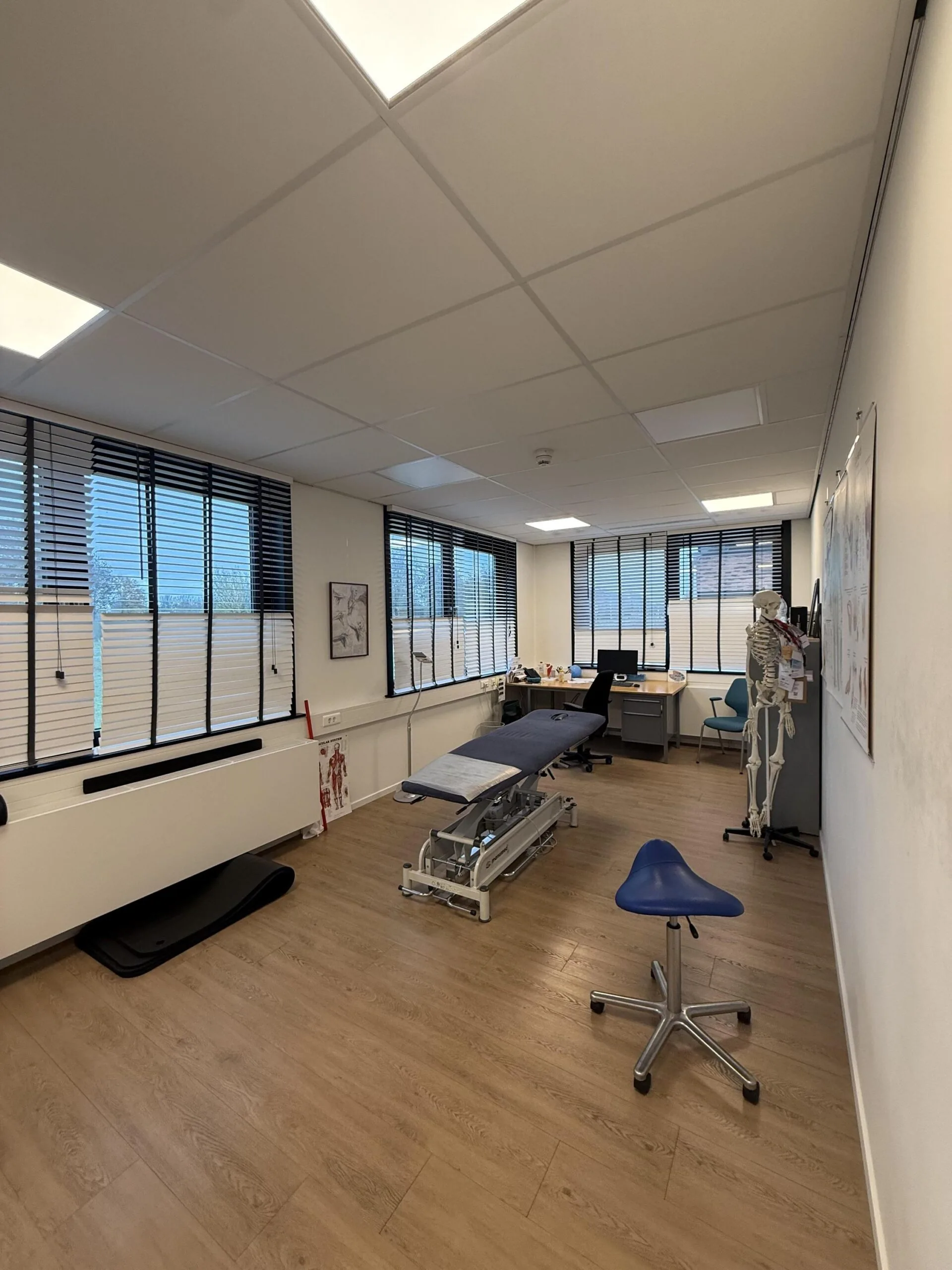 Medical treatment room on the Veteranenlaan with an examination table, anatomical model, and desk near large windows with blinds.